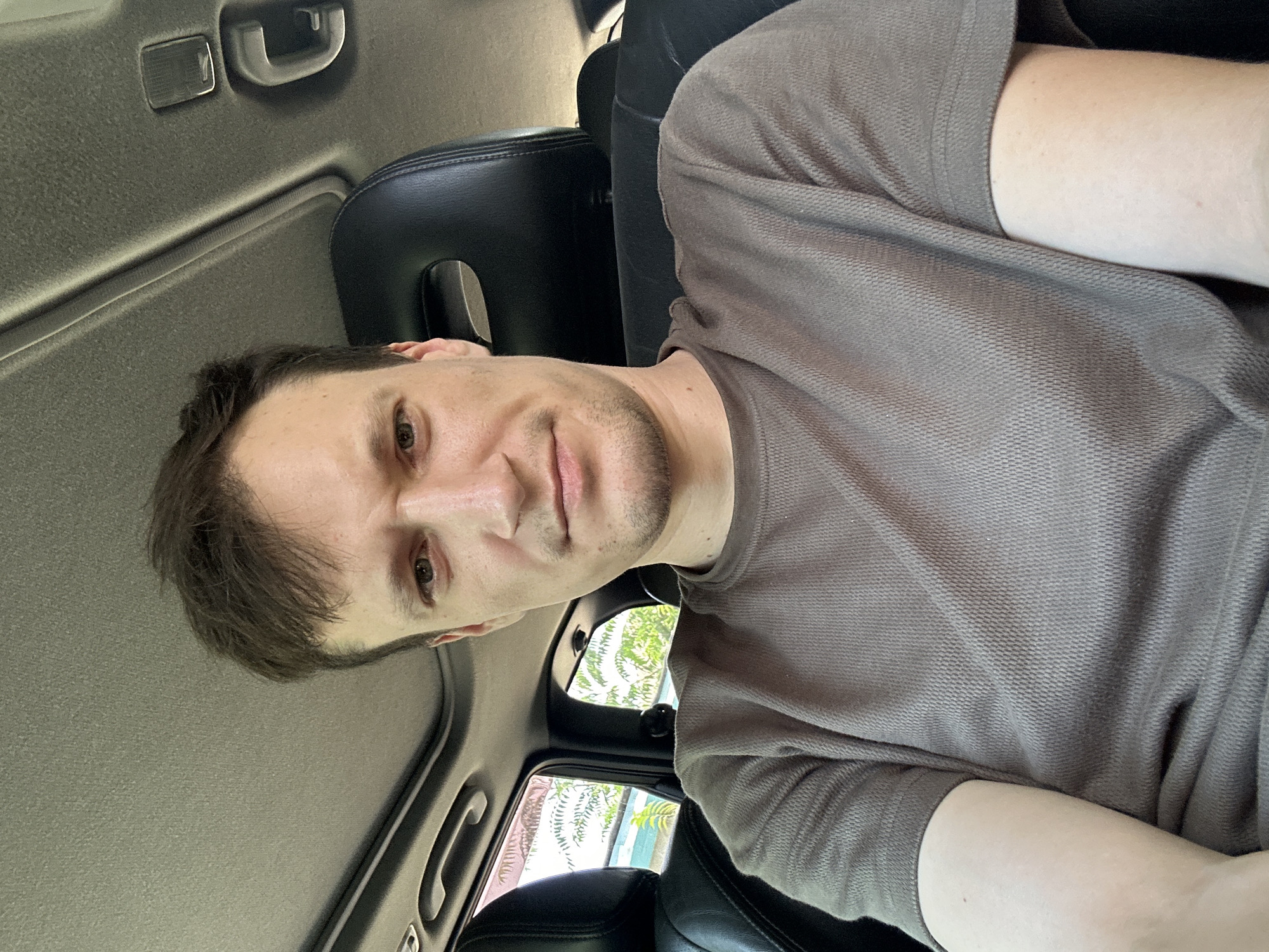 A man is seated in the front passenger seat of a vehicle, looking directly at the camera. The vehicle appears to be parked, with foliage and some buildings visible through the windows. The man is wearing a brown t-shirt and has short brown hair. The interior of the car shows black leather seats and a sunroof. The specific location in the city of Makiv, Ukraine, cannot be determined from the image alone.