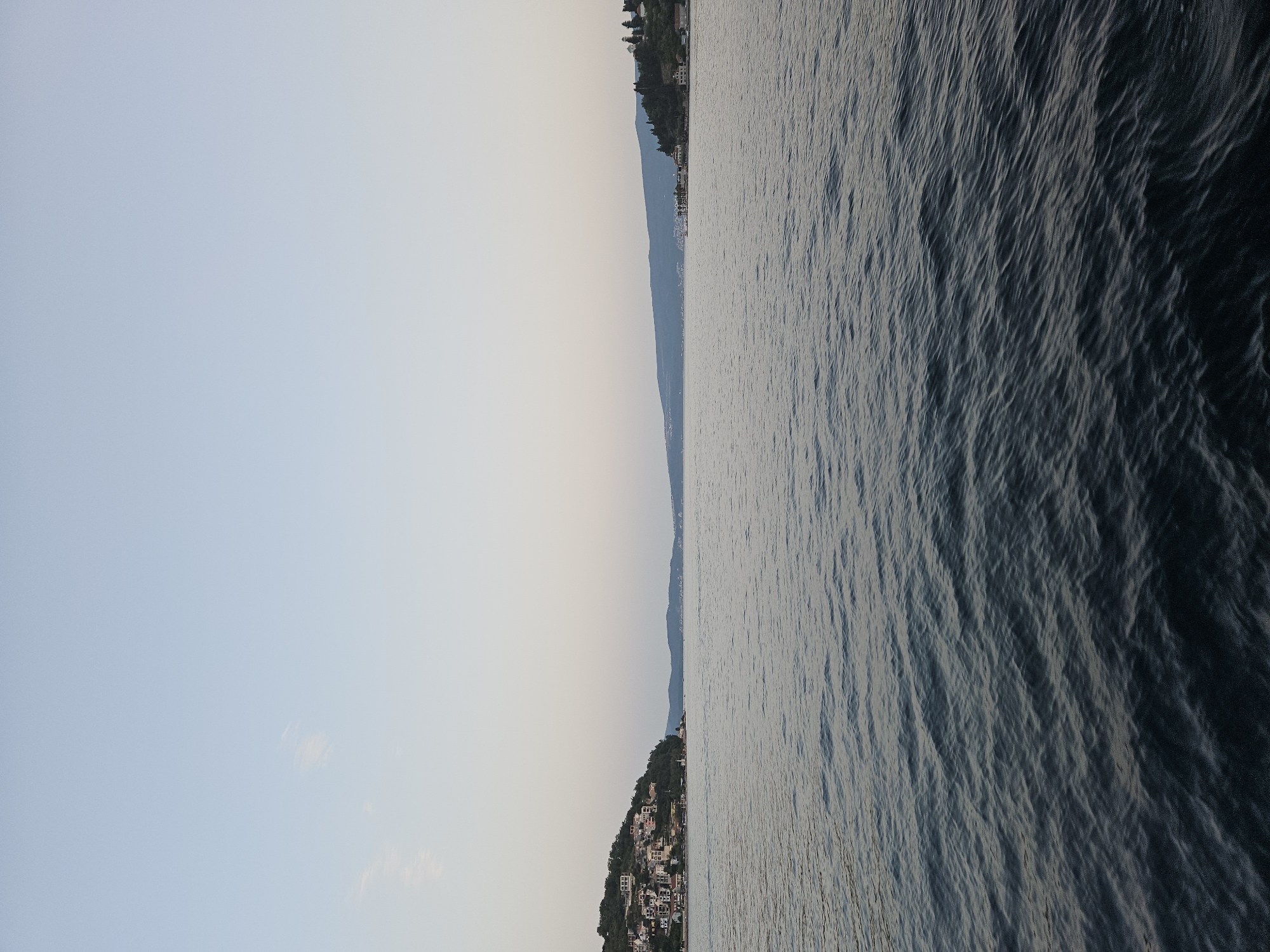 The image shows a body of water with a calm surface, reflecting the pale sky. On the left side, a hillside is densely covered with buildings, suggesting a settlement. The architecture appears to be traditional, with varied roof colors and styles. Trees and vegetation are interspersed among the houses. Further to the left, a coastline curves out of view. On the right side of the image, a similar, though less populated, hillside is visible, also with buildings and trees. The distant horizon is marked by a low-lying landmass, possibly hills or mountains, under a clear, light blue sky that transitions to a lighter hue near the horizon. The water in the foreground is dark blue and shows subtle ripples. The scene evokes a sense of peacefulness and a coastal town setting, consistent with the location of Lepetane, Montenegro.