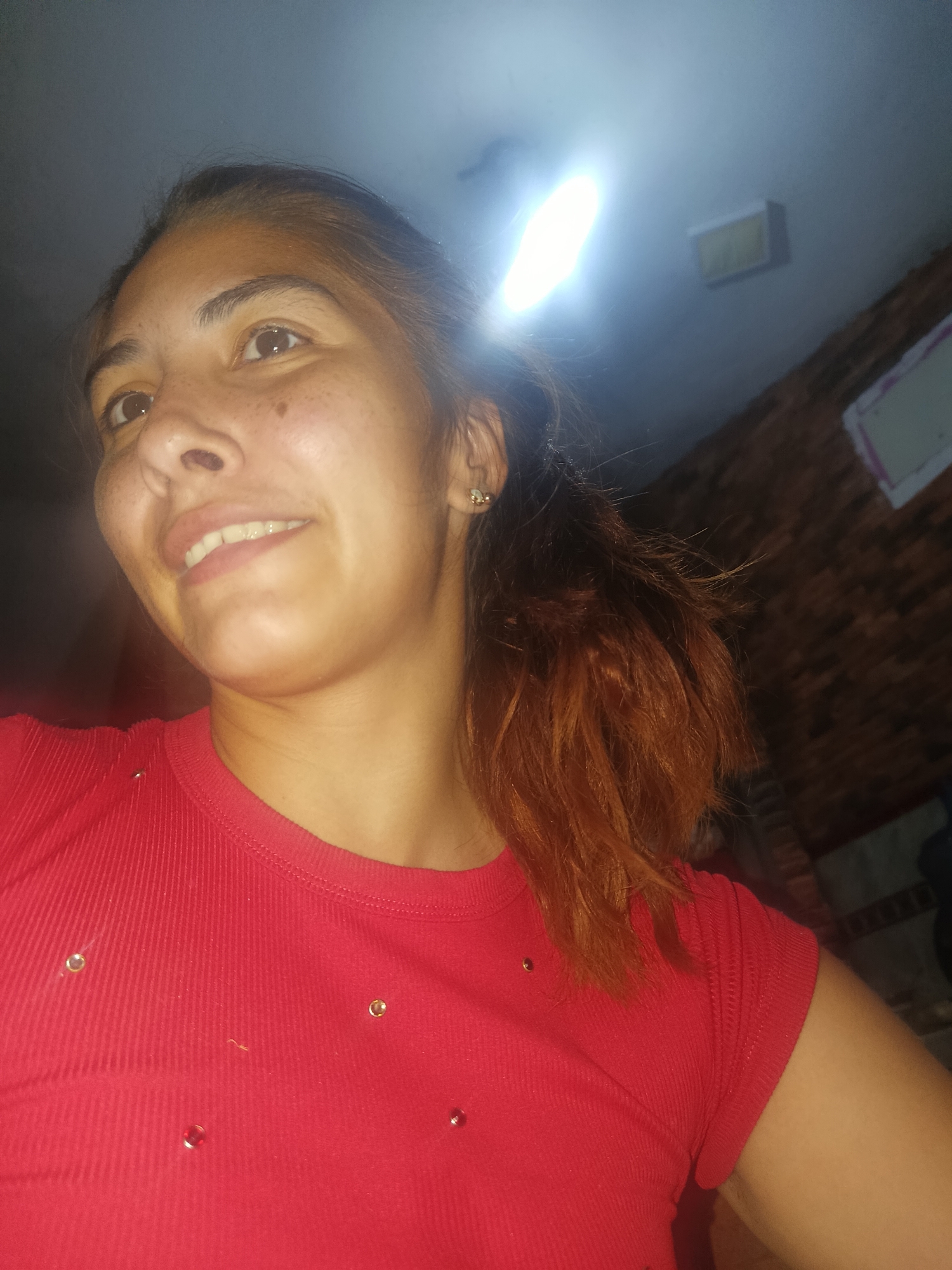 A young woman is captured in a close-up, low-angle indoor shot, looking upwards and to her left with a subtle, gentle smile. She has light brown skin, brown eyes, and dark hair with noticeable reddish-brown highlights or dyed ends, styled in a ponytail. Her face shows a few freckles or moles. She is wearing a bright red, ribbed short-sleeved t-shirt adorned with small, sparkling embellishments scattered across the front. A small, light-colored stud earring is visible in her right ear.
The setting is an indoor location in Juan Griego, Venezuela, likely during the evening or night, as artificial light is the primary illumination source. A very bright, rectangular overhead light fixture, possibly fluorescent or LED, creates a strong glare on the light-colored ceiling directly above her. The background to her right is dark and indistinct, showing a textured wall, possibly brick, and the faint outline of another rectangular object on it. The overall atmosphere is casual, with the woman appearing thoughtful, observant, or pleasantly engaged. No visible text is present in the image.