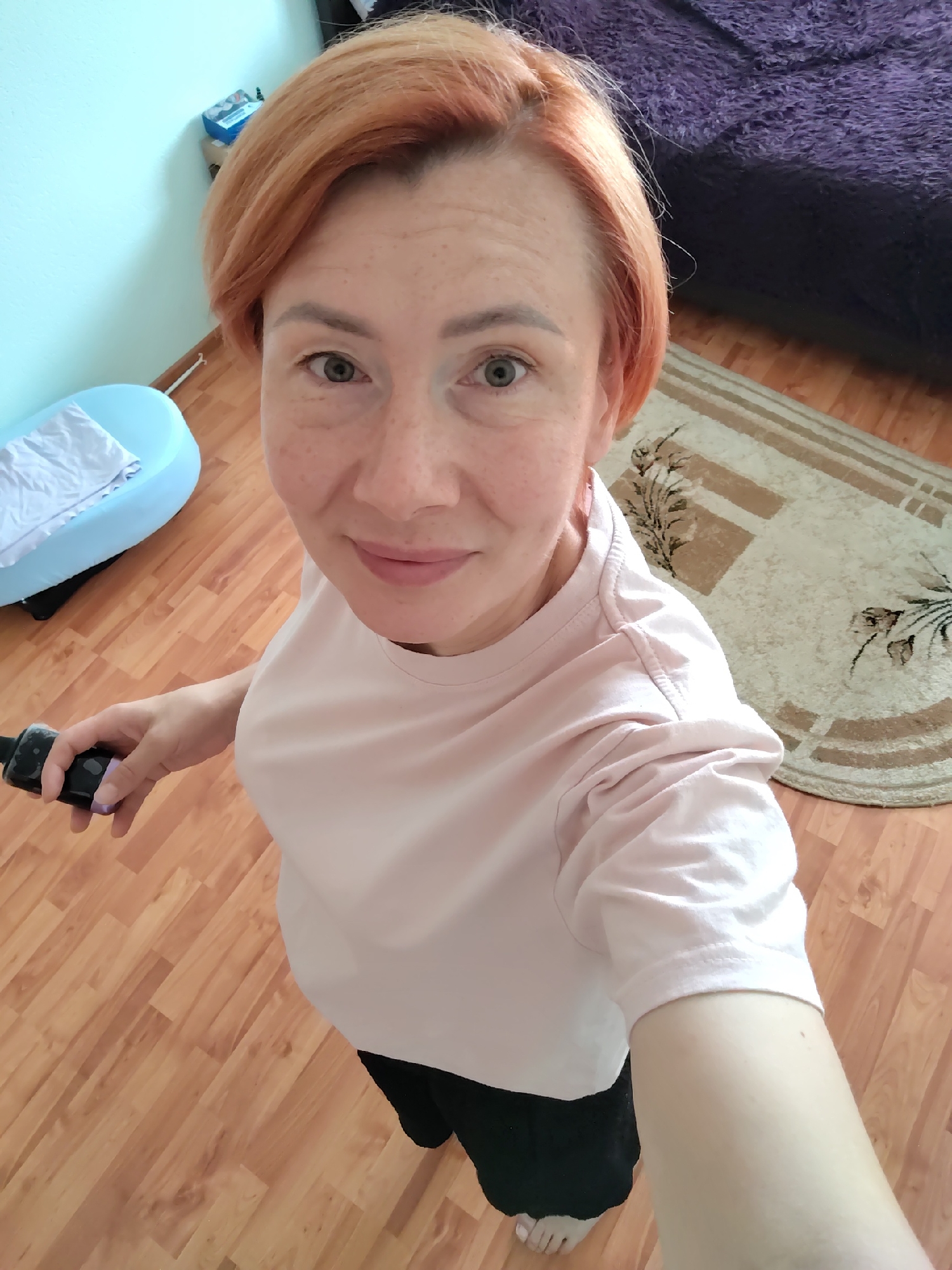 In a residential setting, a woman with short, reddish-blonde hair and freckles takes a selfie. She is wearing a pale pink t-shirt and black bottoms. She holds a black object, possibly a remote or a phone, in her right hand. The background shows a wooden floor, a light blue baby bathtub with a white towel, a rug with a floral pattern, and a dark purple furry surface, likely a bed or sofa. The walls are painted a light blue-green. The overall impression is an indoor domestic scene.