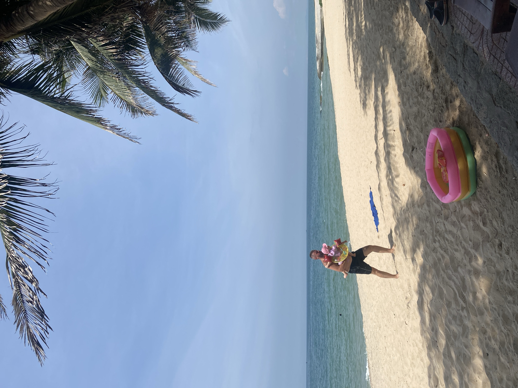 A shirtless man wearing black swim trunks carries a baby on a sandy beach in Phu Thuy Ward, Vietnam. The man is holding the baby with his left arm, and the baby is wearing a pink hat and a yellow and pink float. The beach is wide with light-colored sand, and the ocean is a calm blue. Palm fronds are visible in the upper part of the image, casting shadows on the sand. A small, inflatable kiddie pool with pink and yellow sections is on the sand near the foreground, and a pair of dark shoes are visible to the right. The sky is clear and blue.