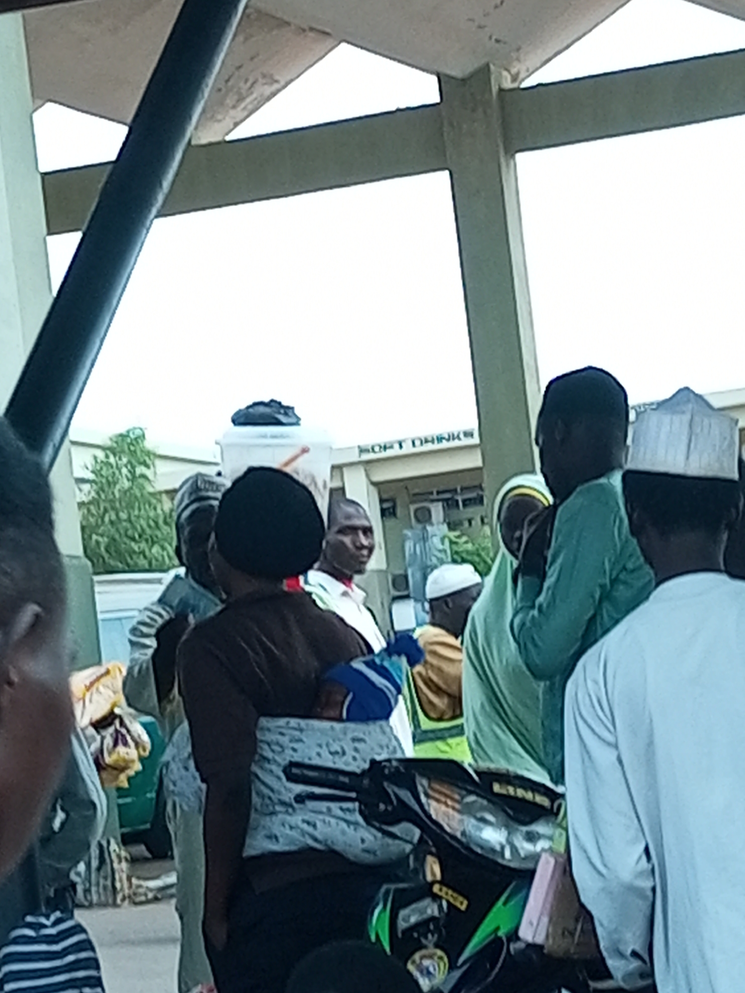 The image depicts a scene in the city of Gombe, Nigeria, likely near a market or transportation hub. Several individuals are present, some engaged in conversation or waiting. A prominent motorbike is visible in the foreground, its headlight illuminated. The background features a building with signs, one of which reads "SOFT DRINKS." The overall atmosphere appears to be that of a bustling public space, with people carrying bags and dressed in casual attire, including headwear common in the region. The lighting suggests it is daytime.