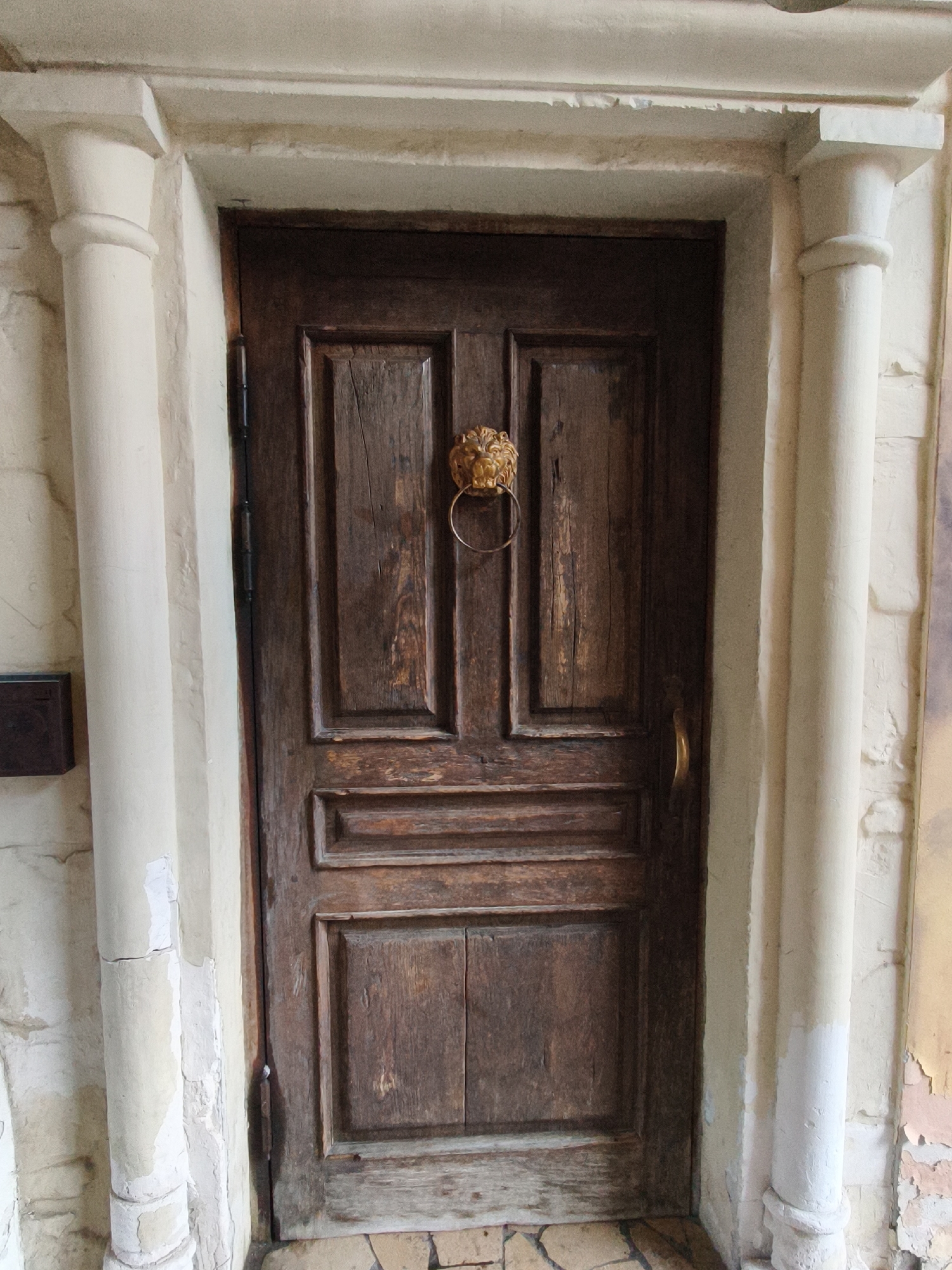 This is a medium-shot, eye-level, portrait photograph of a rustic wooden door with a lion's head knocker. The door is dark brown, with visible grain and signs of age. It is set within a stone archway, flanked by two fluted columns. The columns are a pale, creamy color and appear to be plaster or stucco over stone, with some chipping and wear visible. The doorway itself is framed by a lighter, possibly painted, stone or plaster surround. The wall to the left of the door is textured stone, with a small, dark metal box mounted on it. The wall to the right is lighter in color and also appears to be stone or stucco, with peeling paint in some areas. The floor in front of the door is paved with small, irregularly shaped tiles, some of which are dark and some lighter. The scene appears to be an exterior architectural detail, possibly an entrance to a building or courtyard. The overall impression is one of age and historical character. This photograph was taken in Moscow, Russia.