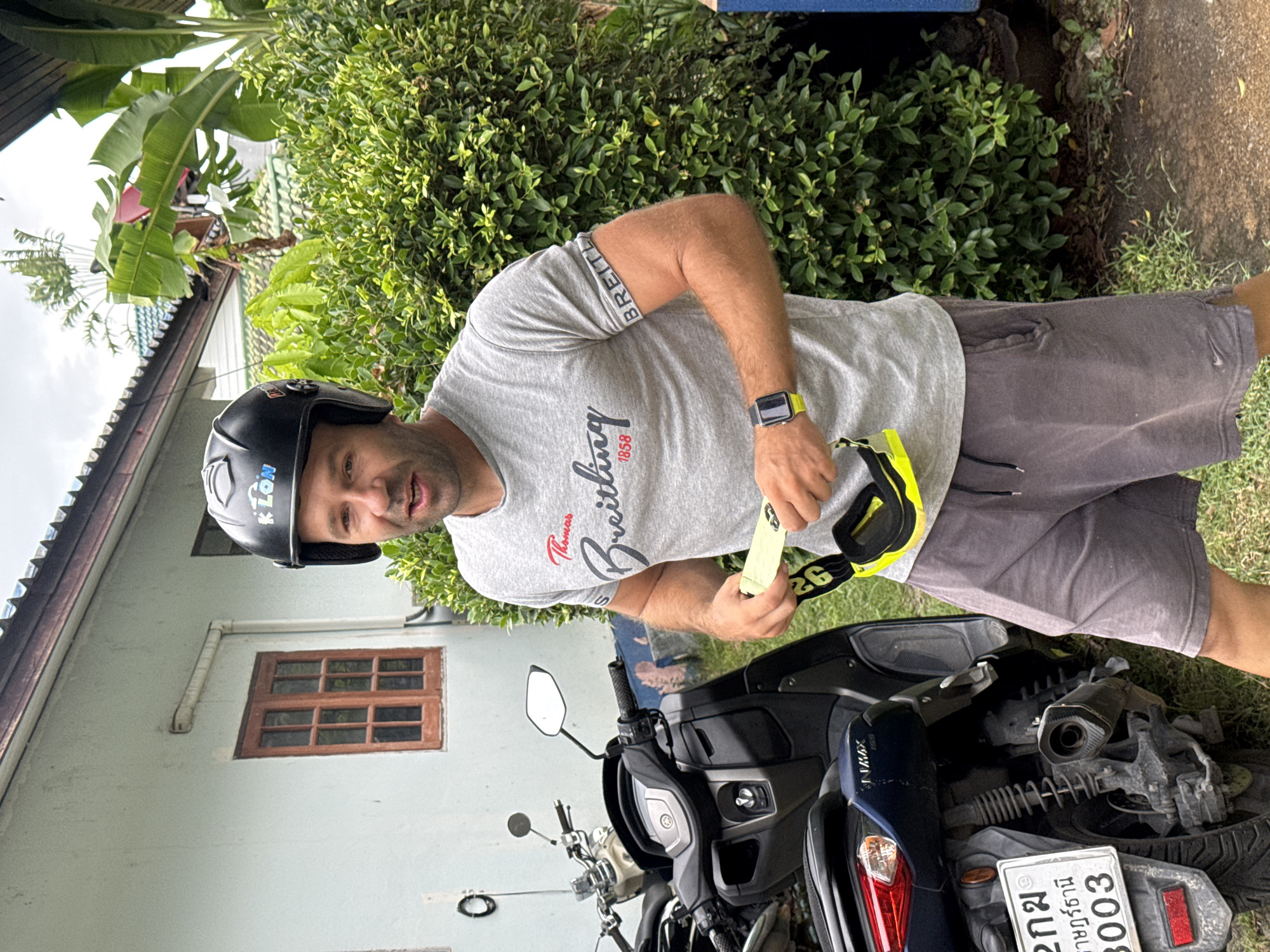 A man wearing a black helmet with "KLON" written on it and a grey t-shirt with "Thomas Breitling 1858" printed on it stands in front of a blue scooter. He is holding a yellow strap with black writing. The man is wearing grey shorts and a smartwatch on his left wrist. The scooter is parked on a paved surface next to a grassy area. In the background, there is a light blue house with a wooden window and a dense green bush. The scene appears to be outdoors in a residential area of Ko Samui District, Thailand, indicated by the license plate on the scooter which contains Thai characters and the number "3003."