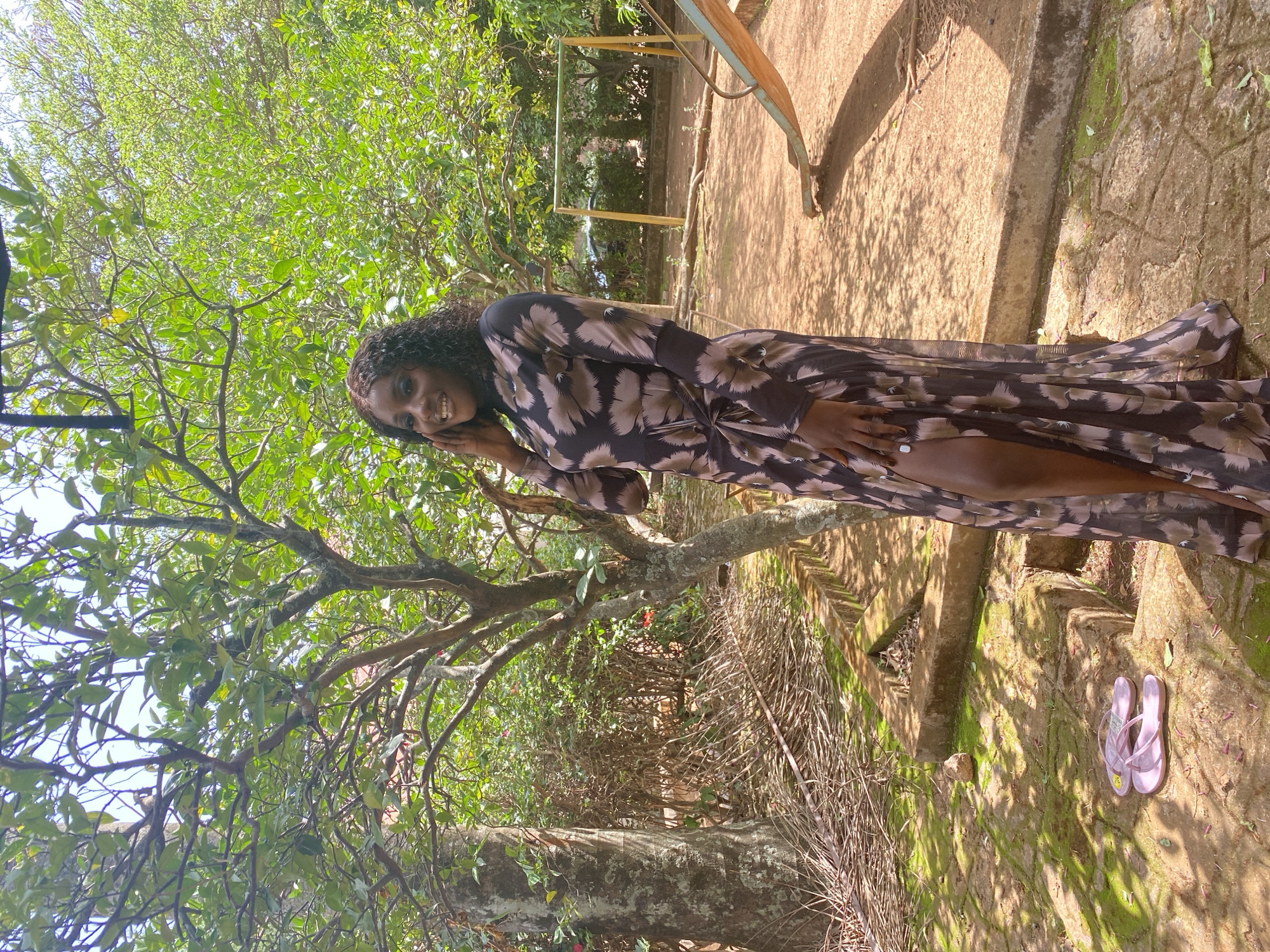 A woman with curly black hair stands outdoors near a tree, smiling with her hand near her face. She wears a long, patterned dress with a belt. The environment consists of trees with green leaves and a sunlit concrete surface with a ledge and moss. In the background, there is a partially visible playground structure. A pair of pink flip-flops rests on the ground nearby. The location is Bukuru, Nigeria.