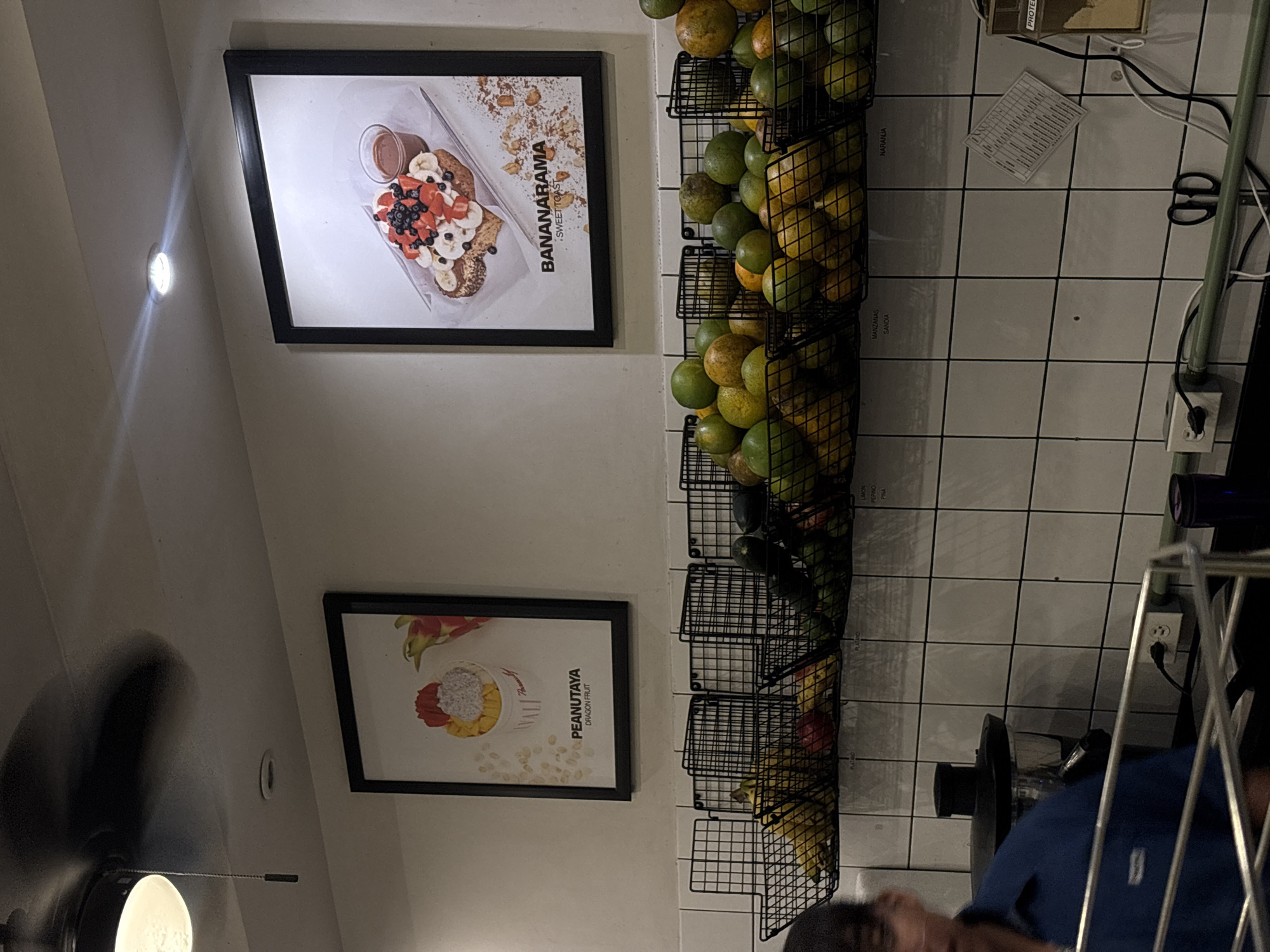 The image shows the interior of a food establishment, likely in Puebla, Mexico. Two framed posters hang on a white wall, one advertising "Bananarama Sweettoast" with a visual of the dish, and the other advertising "Peanutaia Dragon Fruit" with a visual representation. Next to the posters are wire racks stacked with various fruits, potentially for use in food preparation. To the right of the fruit racks is a tiled wall with electrical outlets, cords, and what appears to be a juicer. In the lower right corner, a person wearing a blue shirt is partially visible, suggesting an employee or customer in the establishment. The setting indicates a cafe or restaurant that features fruit-based dishes or smoothies, located in Puebla, Mexico, as evidenced by the name on the framed poster "Bananarama Sweettoast".