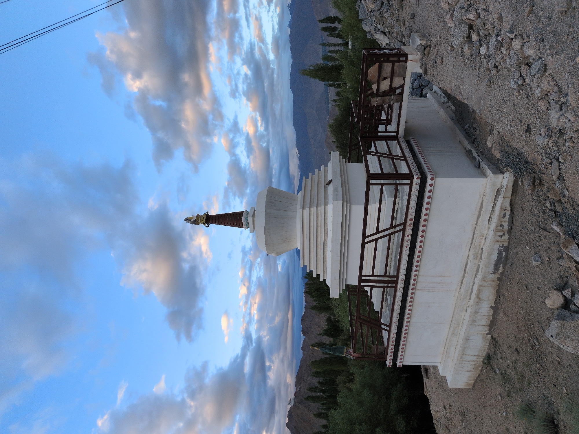 A white stupa stands on a hill in Leh, India, with a backdrop of mountains and a cloudy sky. The stupa, a dome-shaped Buddhist structure, is white with decorative red and white trim around its base. A brown railing with metal supports surrounds a platform leading to the stupa. In the distance, rolling hills are covered with trees and vegetation, and snow-capped mountains rise behind them. The sky is a mix of blue and grey clouds, with hints of pink and orange from the setting sun. The foreground consists of rocky, uneven terrain.