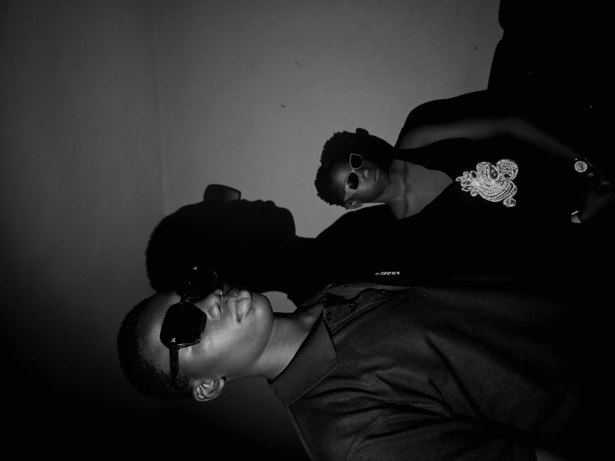 Two individuals are captured in a black and white photograph. The person in the foreground is wearing sunglasses and a collared shirt. Behind them, another individual is wearing sunglasses and a shirt with an ornamental design. The location is Unknown, Unknown. Both people are in front of a plain wall. The lighting casts shadows of the individuals on the wall.