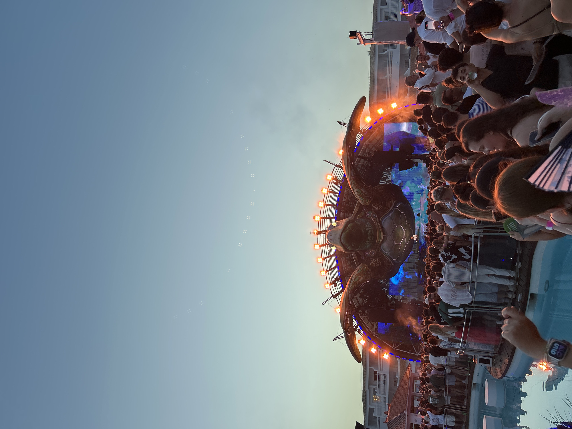 A large crowd of people are gathered at an outdoor event in Sant Josep de sa Talaia, Spain, likely a music festival given the stage setup. The stage features a massive, intricate turtle-shaped structure as the centerpiece, adorned with numerous orange lights. Behind the stage, a multi-story white building is visible, possibly a hotel or resort. The sky above is a clear, pale blue with a gradient effect, and there are small clusters of bubbles scattered across it. The crowd is dense and appears to be enjoying the event, with many holding up their phones. In the foreground, a body of water, possibly a pool, is visible with some round structures along its edge. The overall scene captures a vibrant and energetic atmosphere at dusk.
