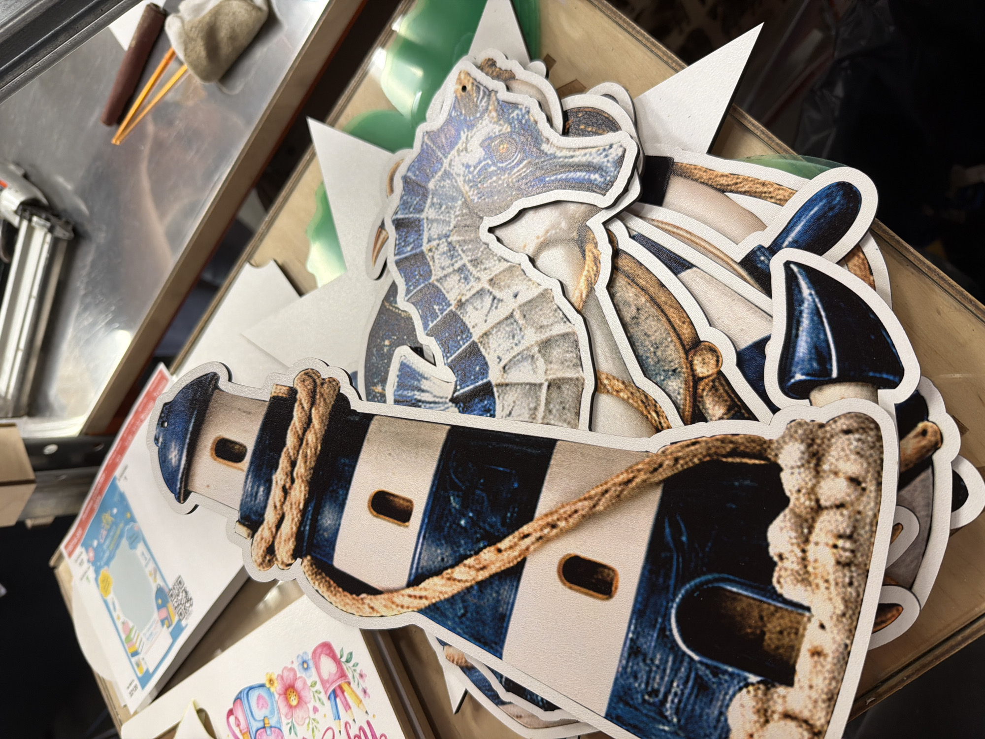 In an indoor setting with ambient lighting, a collection of die-cut paper decorations is arranged on a wooden surface. The primary focus is on several nautical-themed cutouts, including a lighthouse with white and blue stripes, a thick rope coiled around it, and sandy ground at its base. Other cutouts visible include a seahorse with intricate detailing and an anchor. The decorations appear to be layered on top of each other, with some partially obscuring others. In the background, hints of other printed materials and what appears to be a piece of machinery or equipment are present. The overall scene suggests a crafting or preparation area.