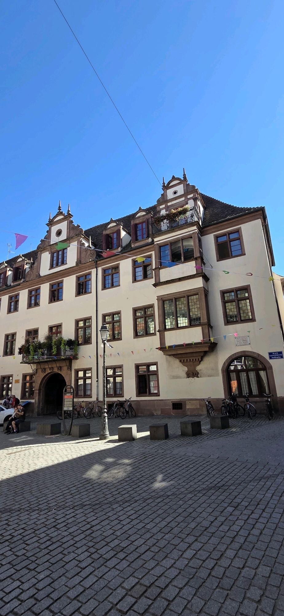 The image depicts a street scene in Strasbourg, France, likely in the historic Petite France district, characterized by its traditional Alsatian architecture. A cream-colored building with a dark brown roof and decorative trim dominates the frame. The building features numerous windows with dark wooden frames and dormer windows on the roof. A balcony with potted plants extends from an upper floor, and colorful triangular flags are strung across the facade. In front of the building, a cobbled street is visible, with several bicycles parked along the side. A lamppost stands in the foreground, casting a shadow on the ground. Two individuals are seated on a low wall to the left, with a car partially visible. The sky is a clear, vibrant blue.
