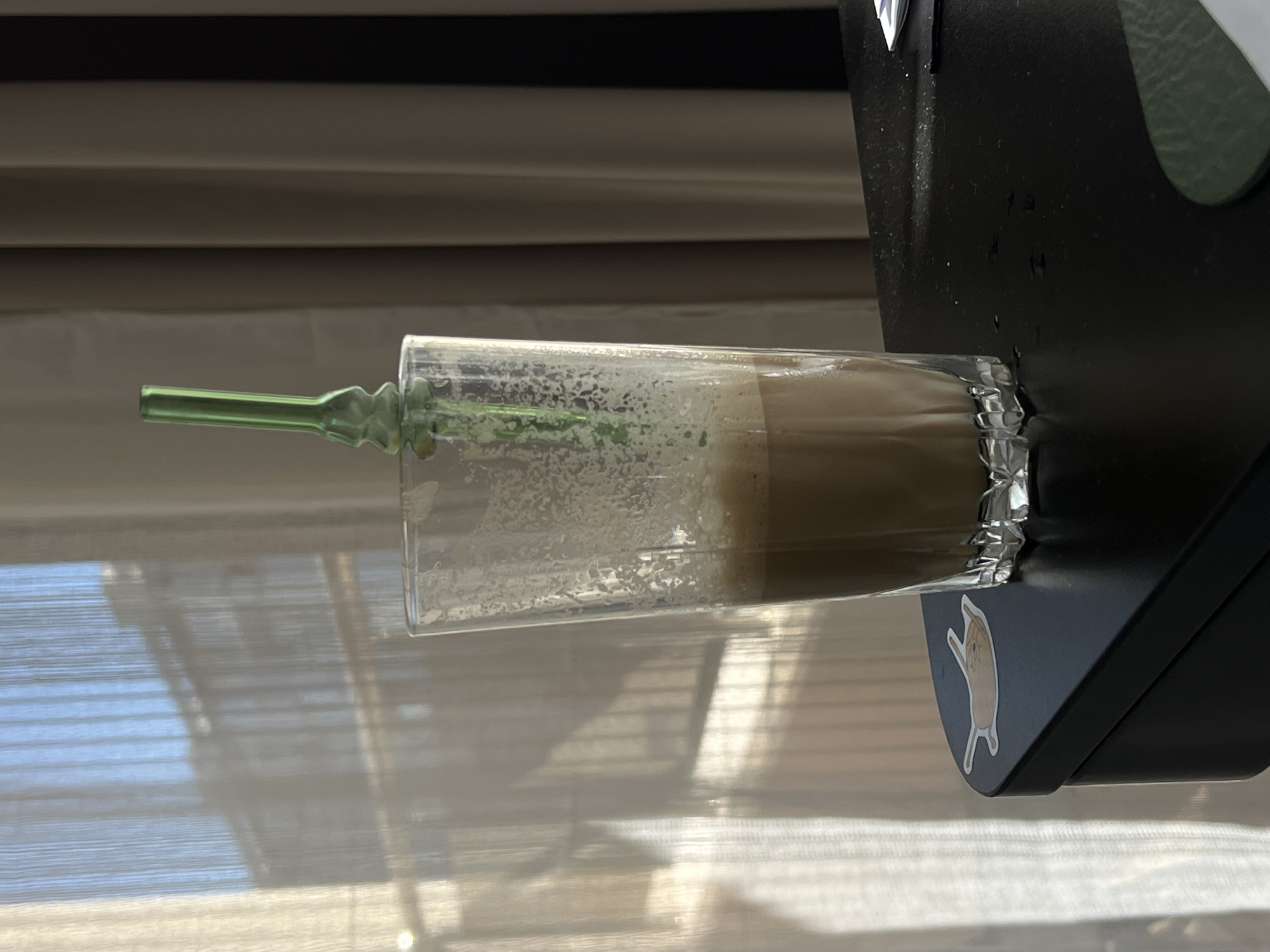 A glass of iced coffee with a green straw sits on a black surface. The surface appears to be a table or desk. Behind the glass, sheer white curtains are visible, with sunlight streaming through them, casting shadows. To the right of the glass, vertical blinds in neutral tones are visible. A small sticker of a cat is on the black surface to the left of the glass. The scene is indoors, likely in a domestic or office setting. There is no identifiable location in Burgas, Bulgaria.