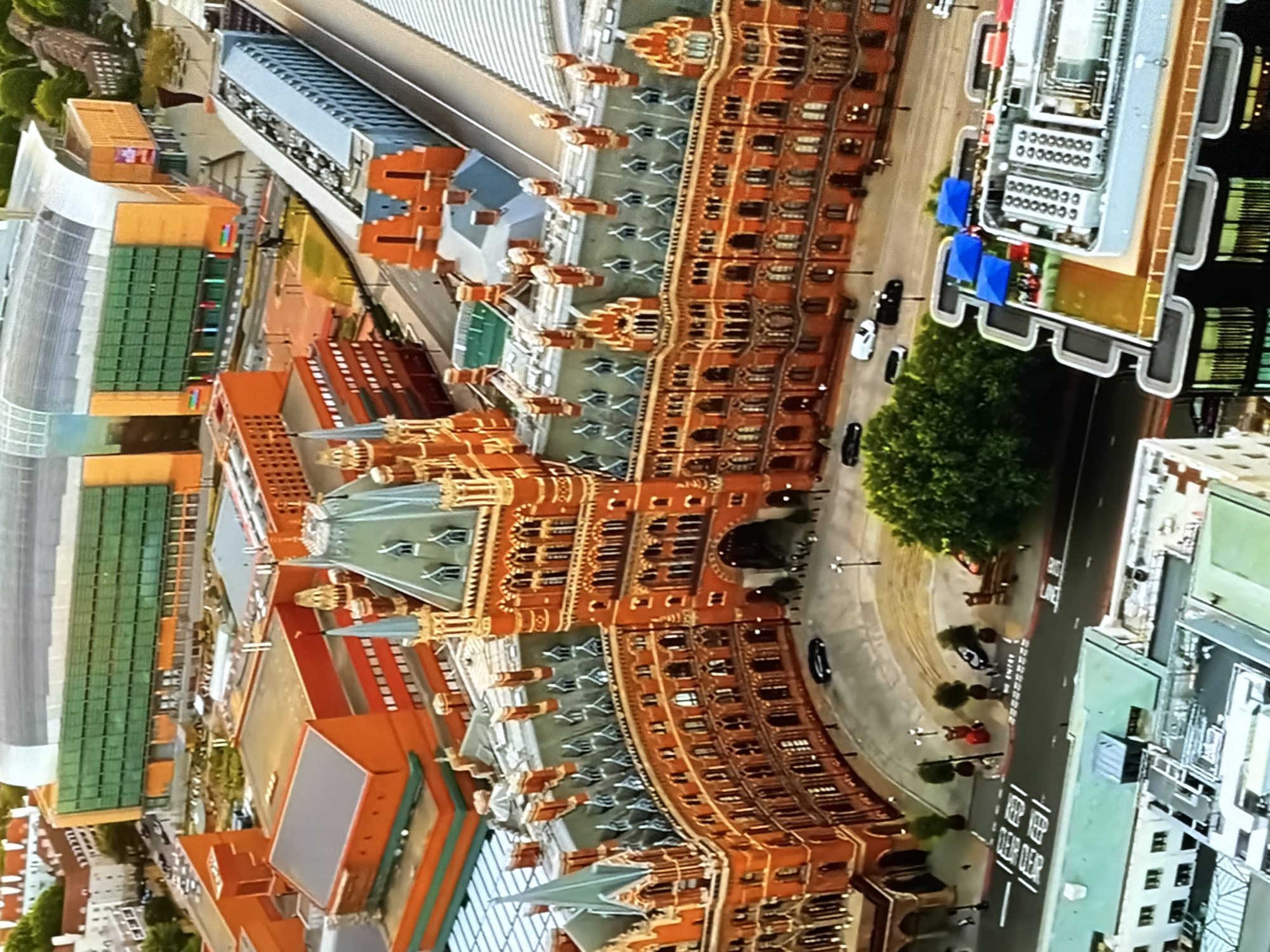 This aerial view captures St Pancras International Station and the St Pancras Renaissance Hotel in London, United Kingdom, under bright daytime conditions. The foreground and middle ground are dominated by the ornate, red-brick Victorian Gothic revival architecture of the St Pancras Renaissance Hotel, featuring multiple spires, turrets, and a complex grey/green roofline. A prominent arched entrance is visible at the center of the hotel's long, curving facade.
In front of the hotel, a curved asphalt road carries several vehicles, including dark cars and a white van, with painted road markings and streetlights. Small figures of pedestrians are visible on the pavements adjacent to the hotel and roads, indicating light activity. A large, lush green tree stands prominently by the roadside in the lower-middle foreground.
To the right of the hotel, multiple railway tracks are visible beneath a modern, curved roof structure, characteristic of a major international station. Further in the background, a large, modern building with a distinctive curved roof and a facade of green and orange/brown panels, identified as the British Library, is prominent. Surrounding these main structures are various other urban buildings, some with flat roofs, and what appears to be a construction or ground works area between the historic hotel and the modern library. On a building in the lower right, a rooftop area with blue canopies and HVAC units is visible.
