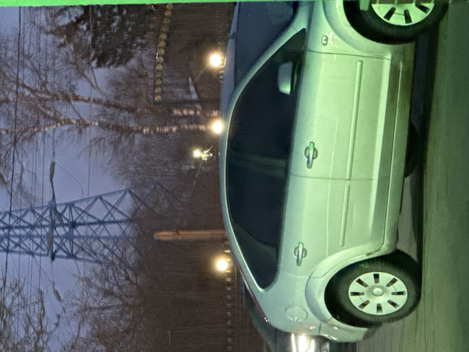 The scene depicts an outdoor setting in 3-Y Uchastok, Russia, under dim, likely evening or night, conditions with a dark blue-grey sky. In the midground, a silver five-door hatchback, consistent with a second-generation Ford Focus, is parked or idling, presenting its left side. Its windows are tinted dark, and the vehicle shows some dirt accumulation on its lower body and wheel wells. Its wheels feature multi-spoke hubcaps. Partial visibility of another vehicle, possibly a darker sedan, is seen to the left and rear of the silver hatchback, with only its front left headlight and bumper visible.
The background features a prominent, light blue or grey metal lattice power transmission tower. Densely packed, bare-branched deciduous trees, including some with distinct white bark, extend across the frame behind the vehicles and the tower, suggesting a cooler season. Several bright, circular yellow-orange light sources are visible within the tree line and above the cars, indicative of streetlights or outdoor lamps. A tall, slender brown vertical pole is positioned behind the silver car. To the far right, an ornate dark metal fence or gate with decorative finials is partially visible, behind which a building structure can be faintly discerned. No people are visible, and no discernible actions or interactions are occurring. The environment presents a mix of natural elements and urban infrastructure.