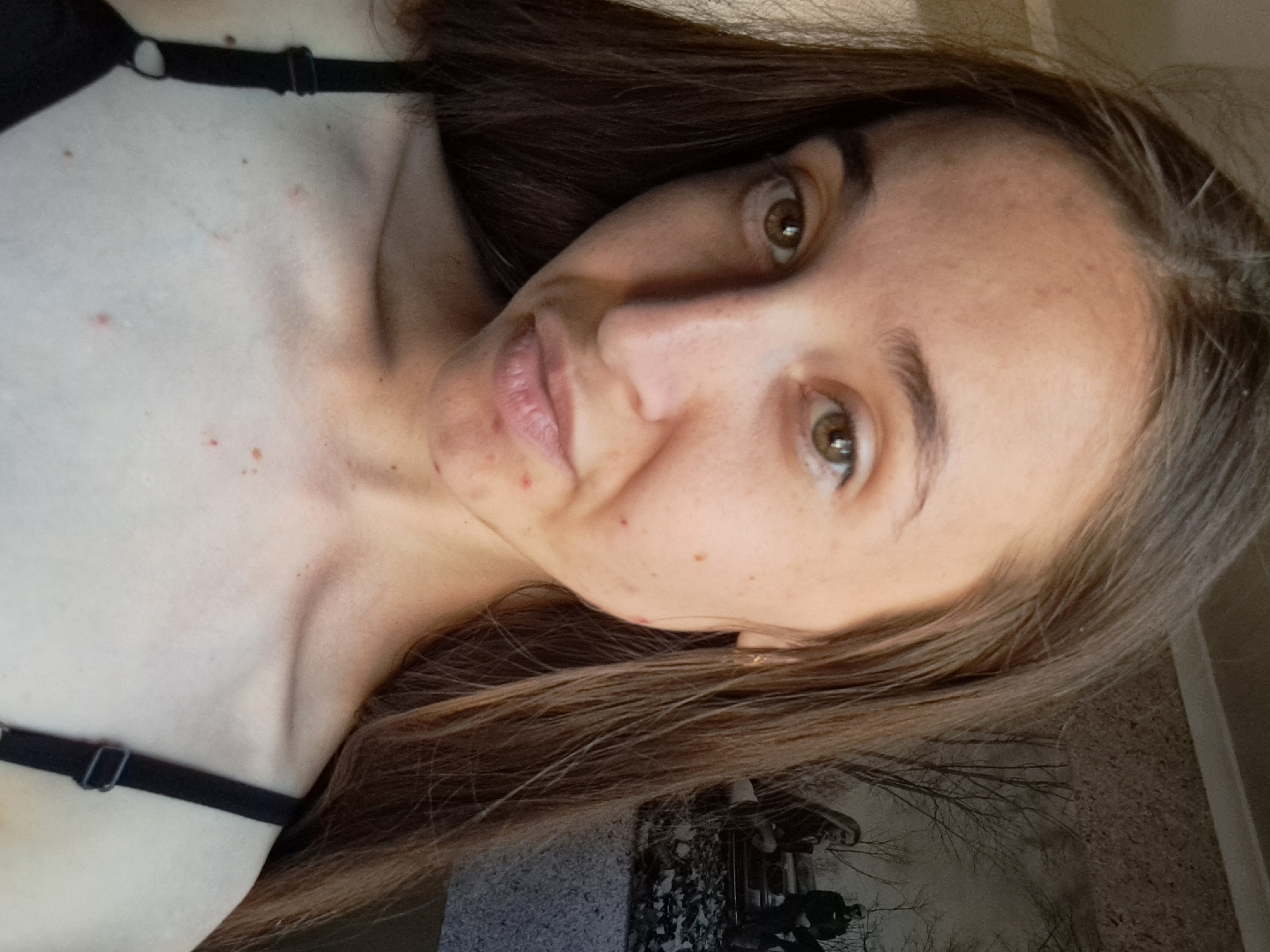 A young woman with long, brown hair is taking a selfie in what appears to be an indoor setting. She is wearing a black bra. The background shows a wallpaper with a scene of trees and people. She has some blemishes on her face and chest. The image is focused on her face and upper body. The location is Vypasne, Ukraine.