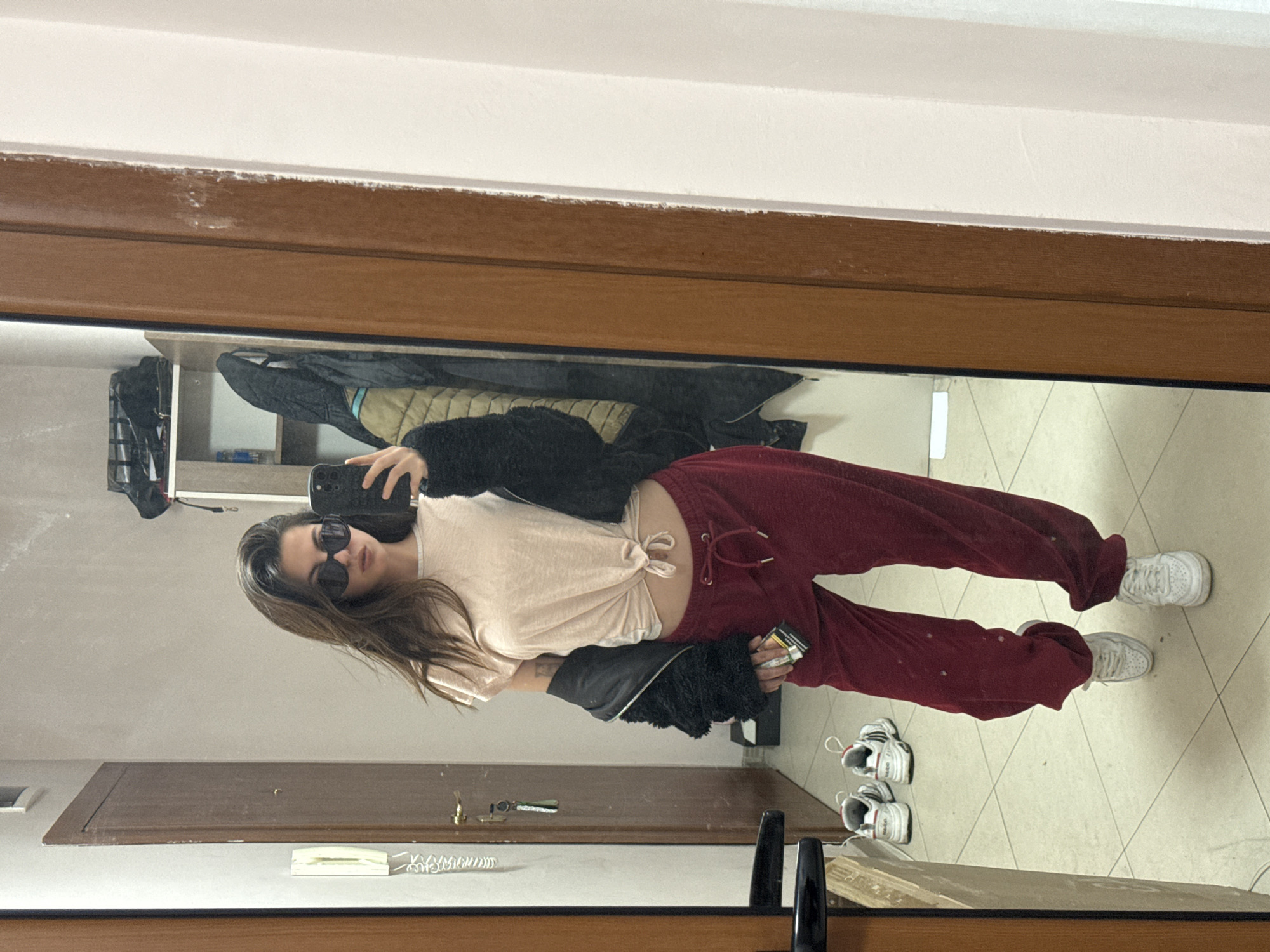A young woman stands in front of a mirror, taking a selfie. She wears sunglasses, a light-colored tied t-shirt, maroon sweatpants, and white sneakers. She holds a smartphone in her left hand and a pack of cigarettes in her right hand. A black jacket hangs on the wall behind her. The reflection in the mirror reveals an interior hallway or entryway with tile flooring, doors, and a shelf with various items. It is not possible to determine if this image was taken in the city of Plovdiv, Bulgaria, or any other specific location.