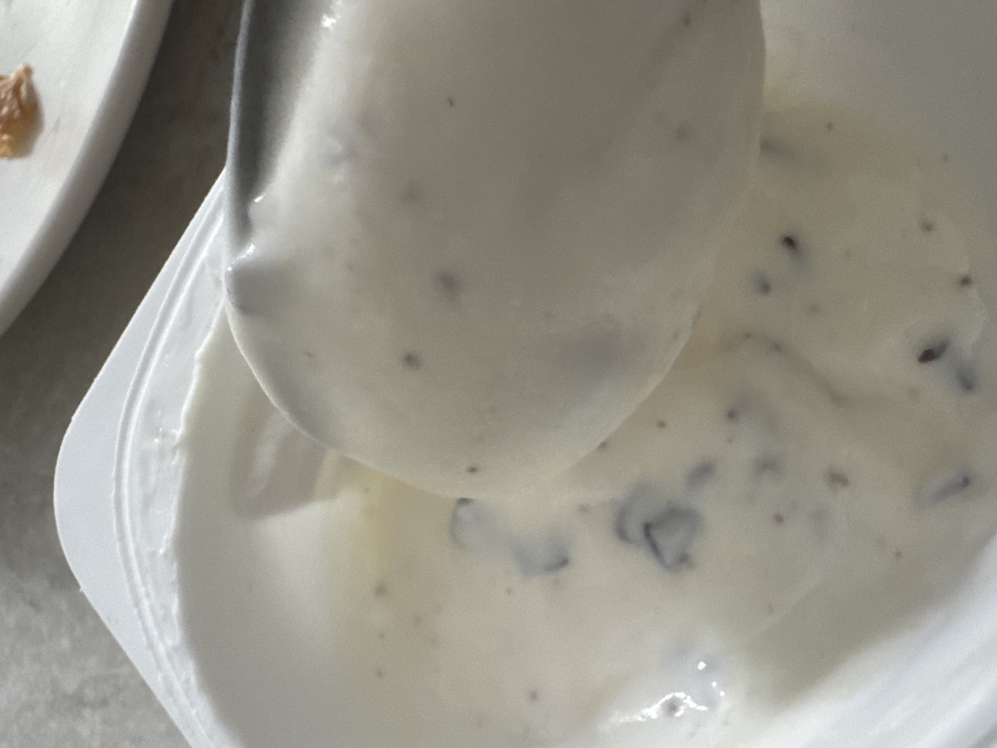 The image shows a close-up view of a spoon scooping white yogurt from a container. The yogurt appears to have small dark specks, possibly fruit or seeds, mixed throughout. A white plastic lid is visible around the edge of the container. The background is out of focus and appears to be a neutral, textured surface, possibly a table or countertop. There is no discernible information about the location in Saratov, Russia, or any people present.