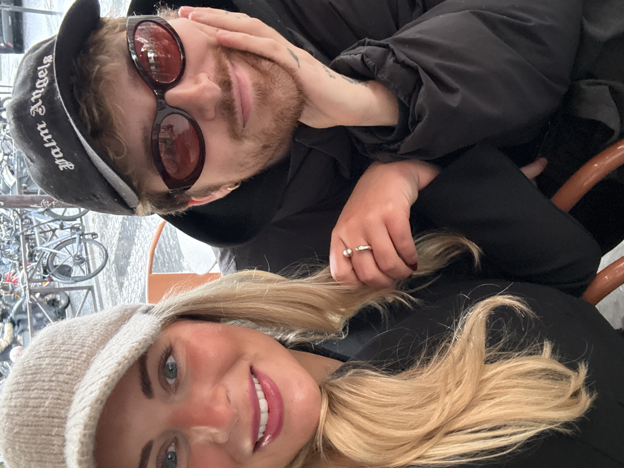 Two people are in a selfie-style photograph rotated 90 degrees clockwise. One person has blond hair and is wearing a knitted beige cap. The other person has light brown hair and a short beard, sunglasses, and a black baseball cap with "Palm Angels" on it. The individuals are wearing dark clothing. The blonde person is smiling. A ring is visible on the hand of one of the individuals. A bicycle rack is visible in the upper left. This could be in Paris, France, because bicycle racks are common in the city.