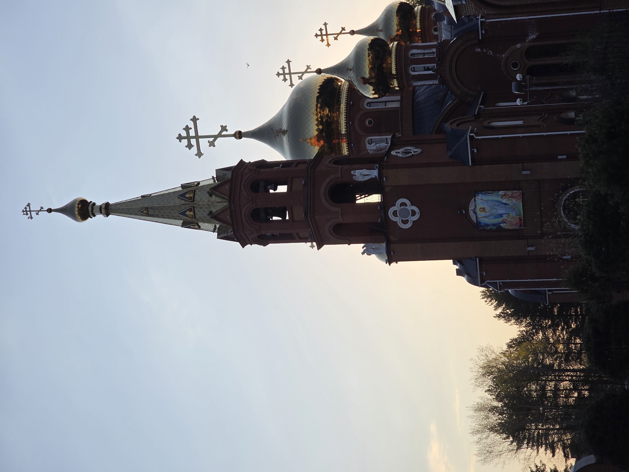 The image depicts a brick Russian Orthodox Church, presumably in Chaikovsky, Russia. The architectural style features multiple domes, each topped with a cross, reflecting traditional design elements. The structure exhibits a mix of reddish-brown brickwork with decorative details and a tall spire with a golden tip. A religious mural can be seen on the side of the building. Trees are visible on the lower right edge of the image, partially obscuring the lower part of the church. The sky is light blue, suggesting either early morning or late afternoon lighting. A bird is also visible in the sky. The photograph is rotated counter-clockwise.