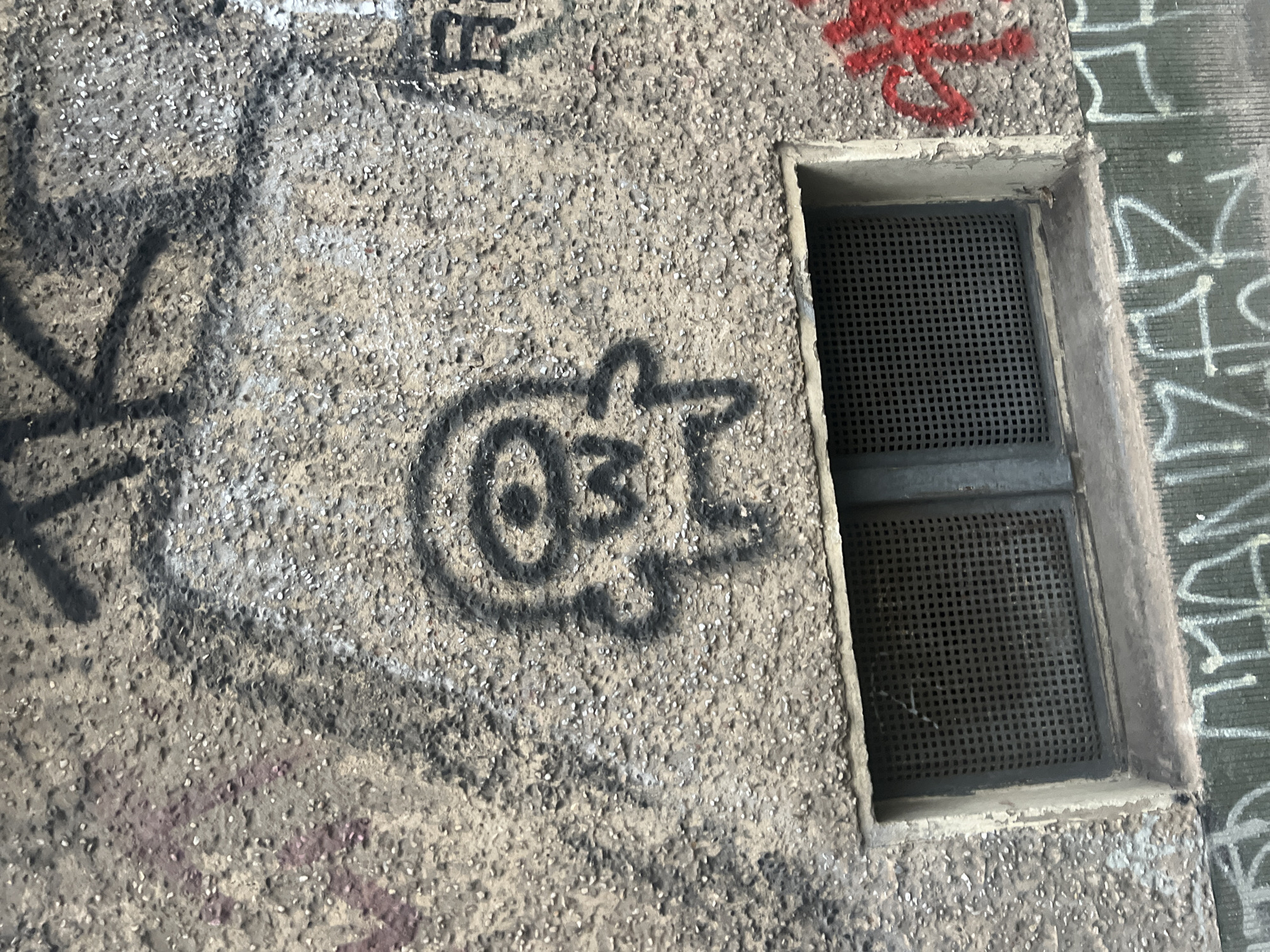 A close-up, eye-level, medium shot captures a textured concrete wall adorned with graffiti. Positioned centrally is a black stencil-style drawing of a cartoonish, cyclopean creature with a wide mouth and triangular teeth. To the left of this figure, faint purple spray paint is visible. Above the creature, dark, thick lines of graffiti suggest letters or symbols. To the right, a partial red graffiti tag is present. Below the central graffiti is a small, recessed window covered with a metal grate. The bottom of the image displays a section of another graffiti tag in white and green. The overall impression is one of urban decay and street art. The graffiti suggests the location is Berlin, Germany.