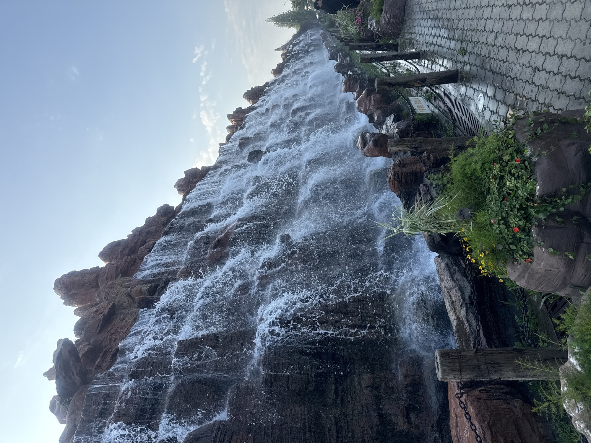 This image depicts a man-made waterfall feature with a rocky, tiered facade. Water cascades down the rocks, creating a dynamic spray. A paved walkway runs alongside the waterfall, separated by a low barrier of rocks and wooden posts connected by chains. Lush green plants and small yellow and red flowers are interspersed among the rocks near the walkway. In the background, a few individuals are visible near the right edge of the frame, and a tall evergreen tree stands further in the distance. The sky is clear blue with a few wispy clouds. This waterfall is located within the Happy Dream World theme park in Qinhuangdao, China.