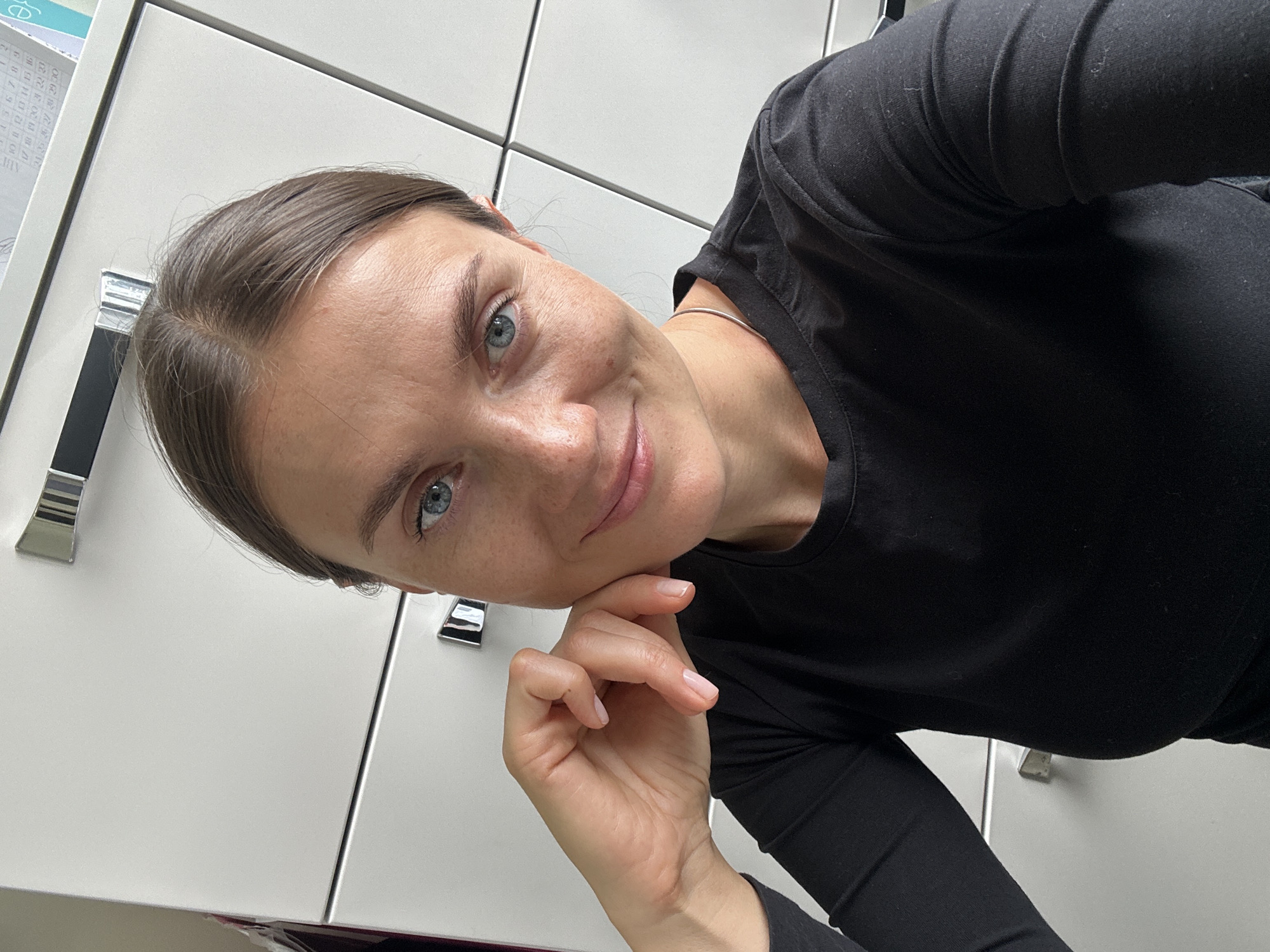 A woman with brown hair and blue eyes, wearing a black long-sleeved shirt, smiles at the camera. Her left hand rests on her cheek, and she is wearing a silver necklace. The background appears to be a white cabinet with silver handles. The image is a selfie taken in an indoor setting. There is no discernible information to indicate the location is Kazan, Russia.