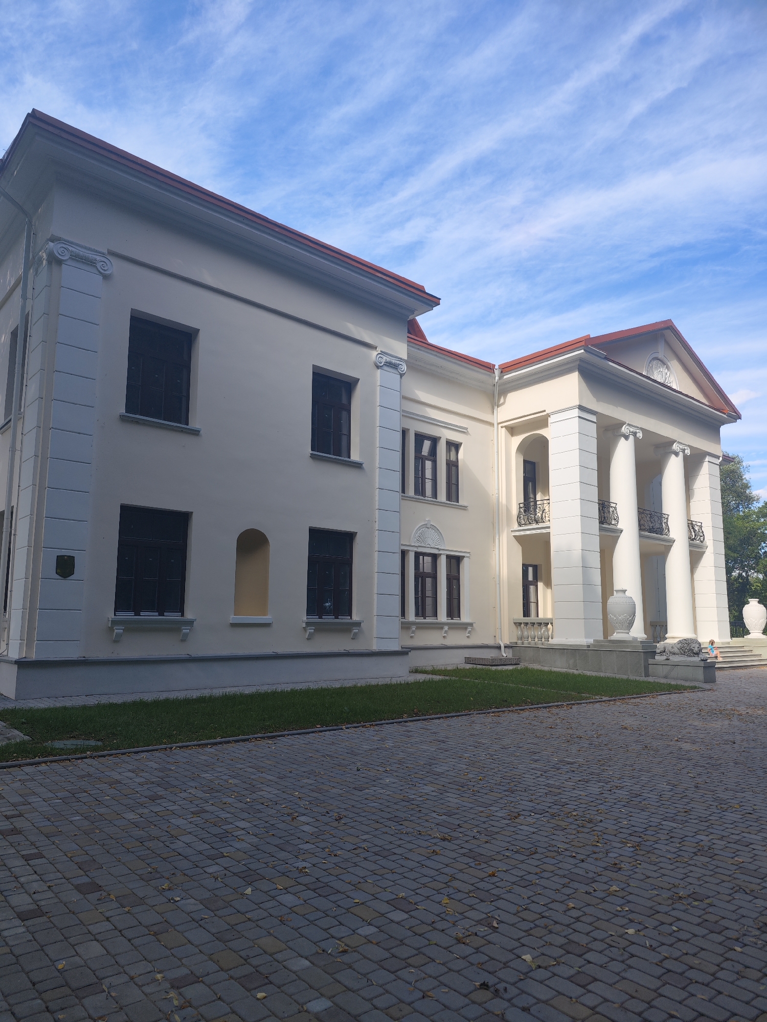 The image shows the facade of a light-colored, multi-story building with classical architectural elements, likely a museum or administrative building, situated in the city of Slonim, Belarus. The building features a prominent portico with large white columns, a pediment with a relief sculpture, and ornate balconies. The exterior walls are decorated with pilasters and cornices. Dark-framed windows are visible on multiple levels. In front of the building, a paved area with interlocking stone pavers leads to the entrance, and a strip of green lawn runs along the base of the structure. Two large, decorative vases and a sculpture of a lion are placed near the entrance. The sky above is blue with wispy white clouds. A small child in a pink outfit is briefly visible on the steps leading up to the portico, interacting with another person who is partially obscured. The scene is captured from a slightly low angle, emphasizing the grandeur of the building.