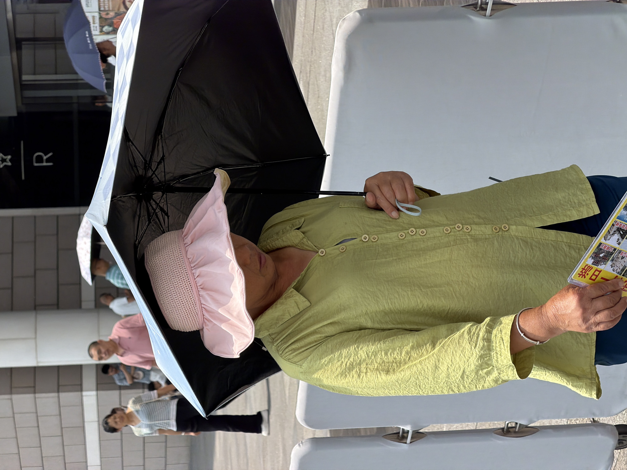An individual wearing a light green, long-sleeved shirt and a pink wide-brimmed hat holds a black umbrella over their head while walking outdoors. The individual is also holding a booklet or pamphlet in their right hand, which features images and text in Chinese. The person wears a silver bracelet on their left wrist. In the background, other pedestrians are visible, some also holding umbrellas, suggesting a sunny or possibly rainy day. The environment appears to be an urban setting, possibly in the Yuzhong District of China, with modern building architecture and paved ground. White barriers are present in the foreground.