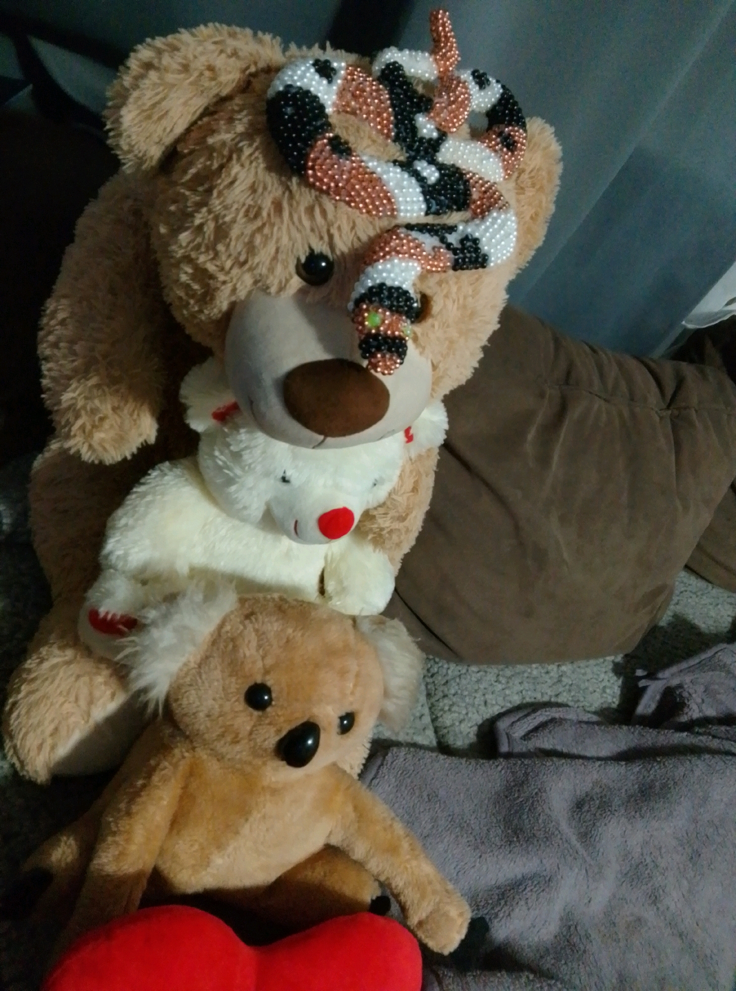 The image shows a collection of stuffed animals. In the foreground, a brown teddy bear is sitting with a red heart-shaped pillow in front of it. Behind this teddy bear, a white stuffed animal, possibly a lamb or a polar bear, is visible. It has a red nose. On top of the white stuffed animal and partially obscuring the face of a large brown teddy bear in the background, is a beaded snake accessory. The large brown teddy bear has a beaded snake wrapped around its head. The stuffed animals are arranged on a grey, textured surface, possibly carpet or a blanket, with some darker fabric in the background. There are no people visible in the image. The location in the city of Wedel, Germany, is not discernable from the visual information provided in the image.