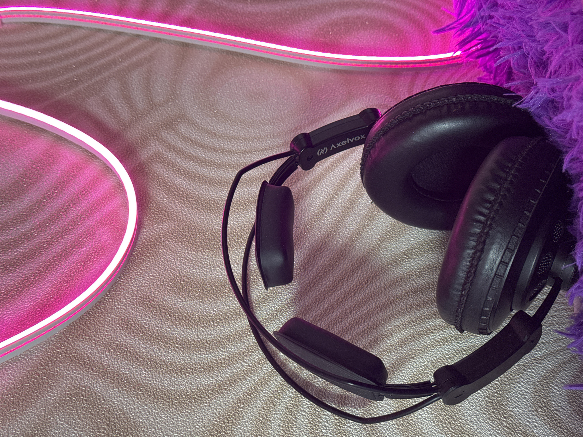 A pair of black over-ear headphones rests on a textured surface, likely a carpet or similar material. Part of a bright pink neon light is visible along the top and left edges of the image, providing illumination. A purple, fluffy object is situated on the right side, partially obscuring the headphones. The headphones are marked "Axelvox" on the side. The image provides no indication of the location being Ново-Никольское, Russia. There are no people visible in the image.