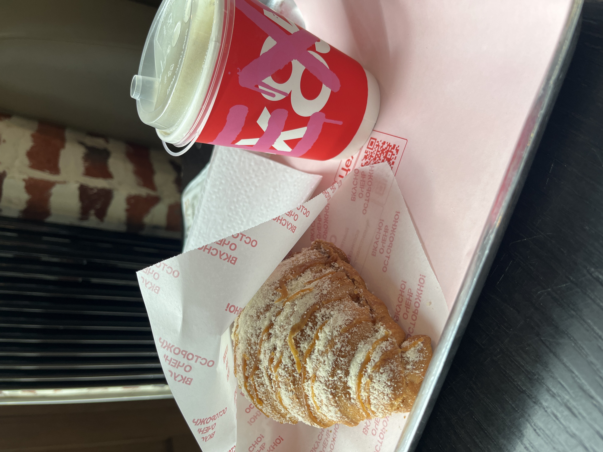 A cup with a lid and a croissant on a branded paper sheet rest on a tray. The cup is red with pink lettering and a clear plastic lid. The croissant is topped with powdered sugar. The paper sheet has red lettering printed on it, and a QR code. The tray is on a surface with dark slats. In the background, a brick wall is partially visible. The branding on the paper sheet and cup suggests a location in Shushary, Russia.