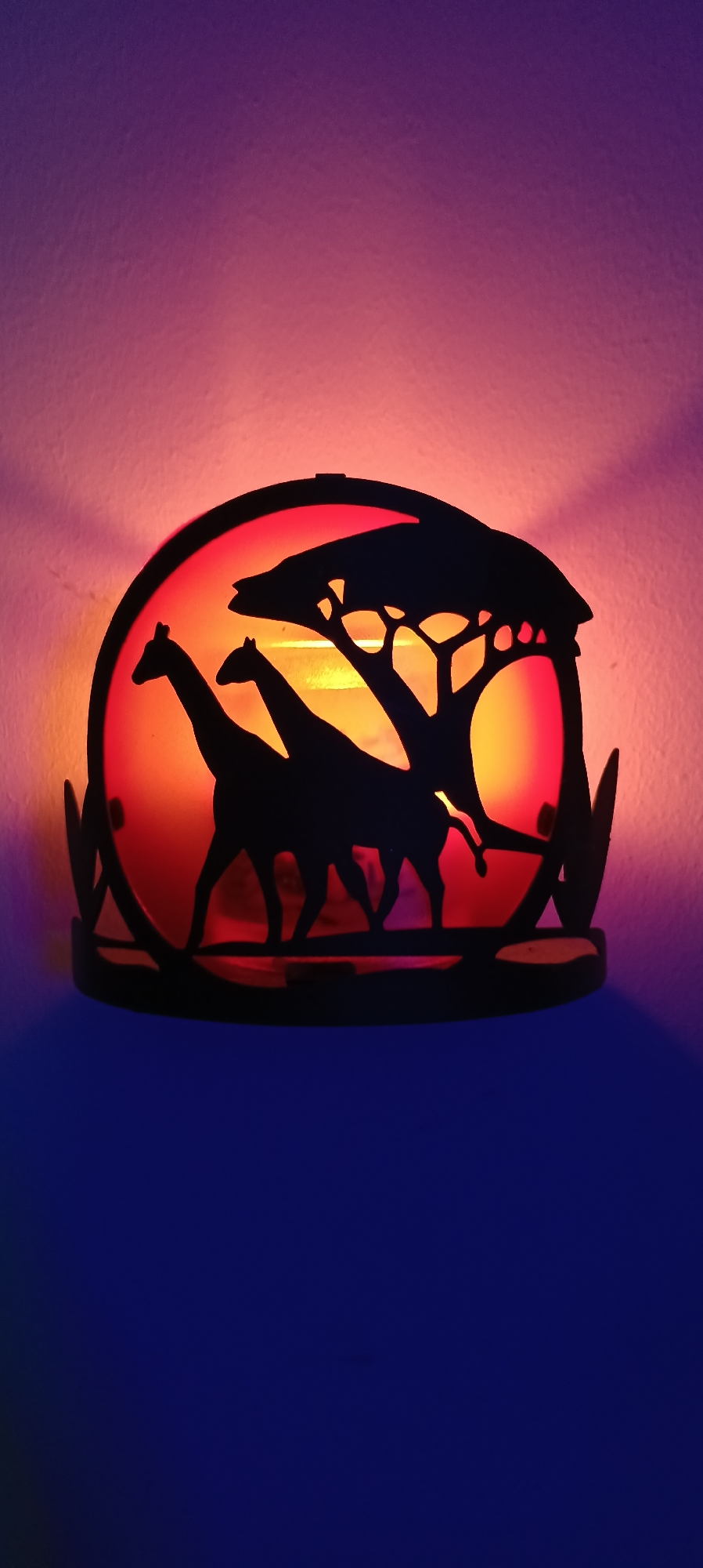 The image shows a silhouette of two giraffes and an acacia tree against a warm, illuminated background. The silhouette is made of dark metal and is framed by a decorative metal border with leaf-like elements on the sides. The background appears to be a light source, possibly a candle or lamp, casting a gradient of red, orange, and yellow light. The overall scene is set against a textured wall that transitions from purple at the top to a deep blue at the bottom, suggesting ambient lighting or a colored backdrop. The object is likely a decorative lamp or a shadow caster. The location in the city of Wels, Austria, is not discernible from the image itself.