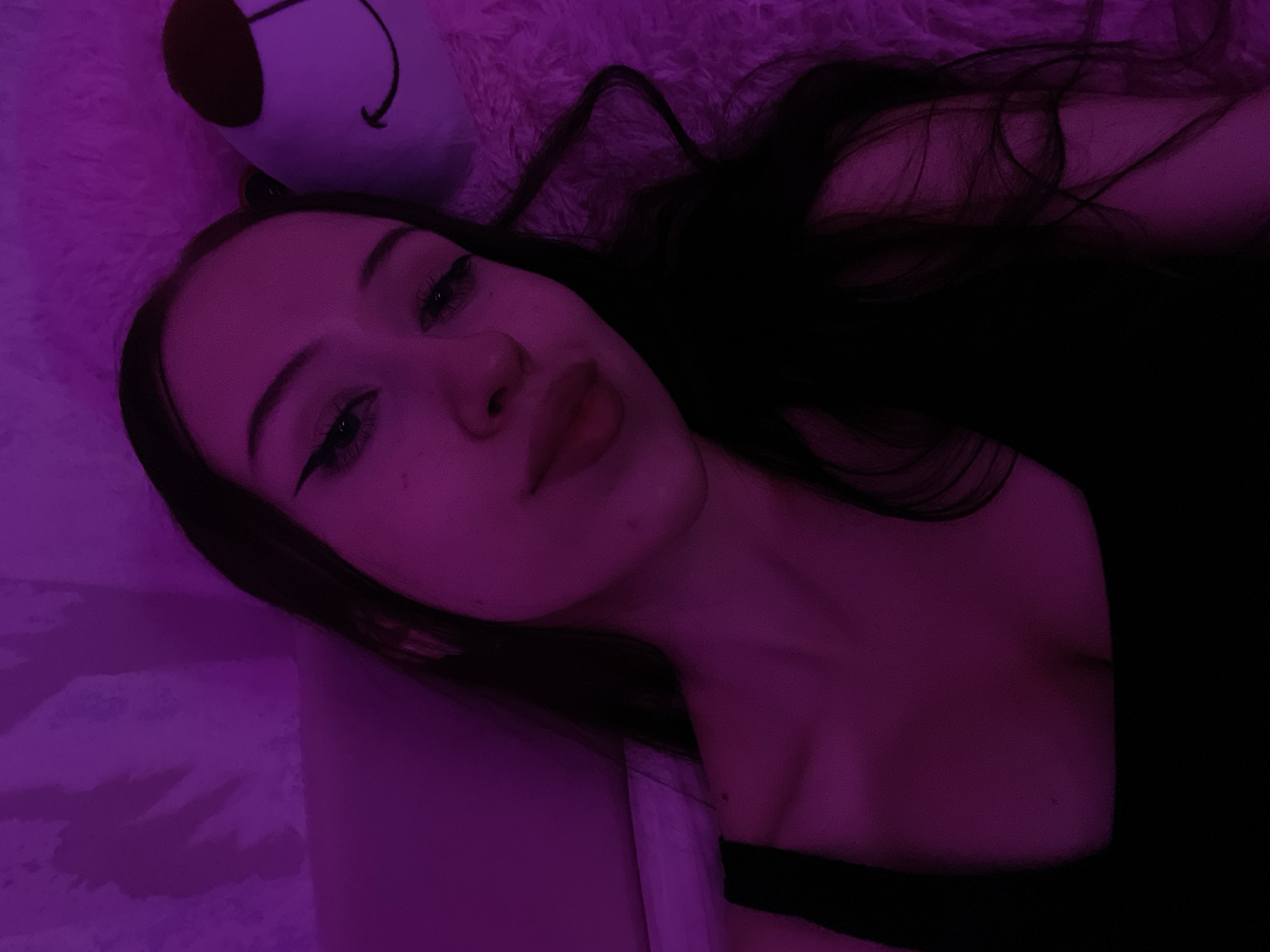 A young woman with long, dark hair, wearing a black top, is lying down. She is looking at the camera with her head tilted. She has on winged eyeliner and her lips are slightly parted. To her right, partially visible, is a plush toy that appears to be a bear with brown ears and a smiling face. The background is a soft, fuzzy pink material, and there is a wall behind her with some textured shadows. The scene is illuminated by purple light, casting a pinkish-purple hue over everything. The image does not provide any information that would allow for the identification of a specific location in Votkinsk, Russia.