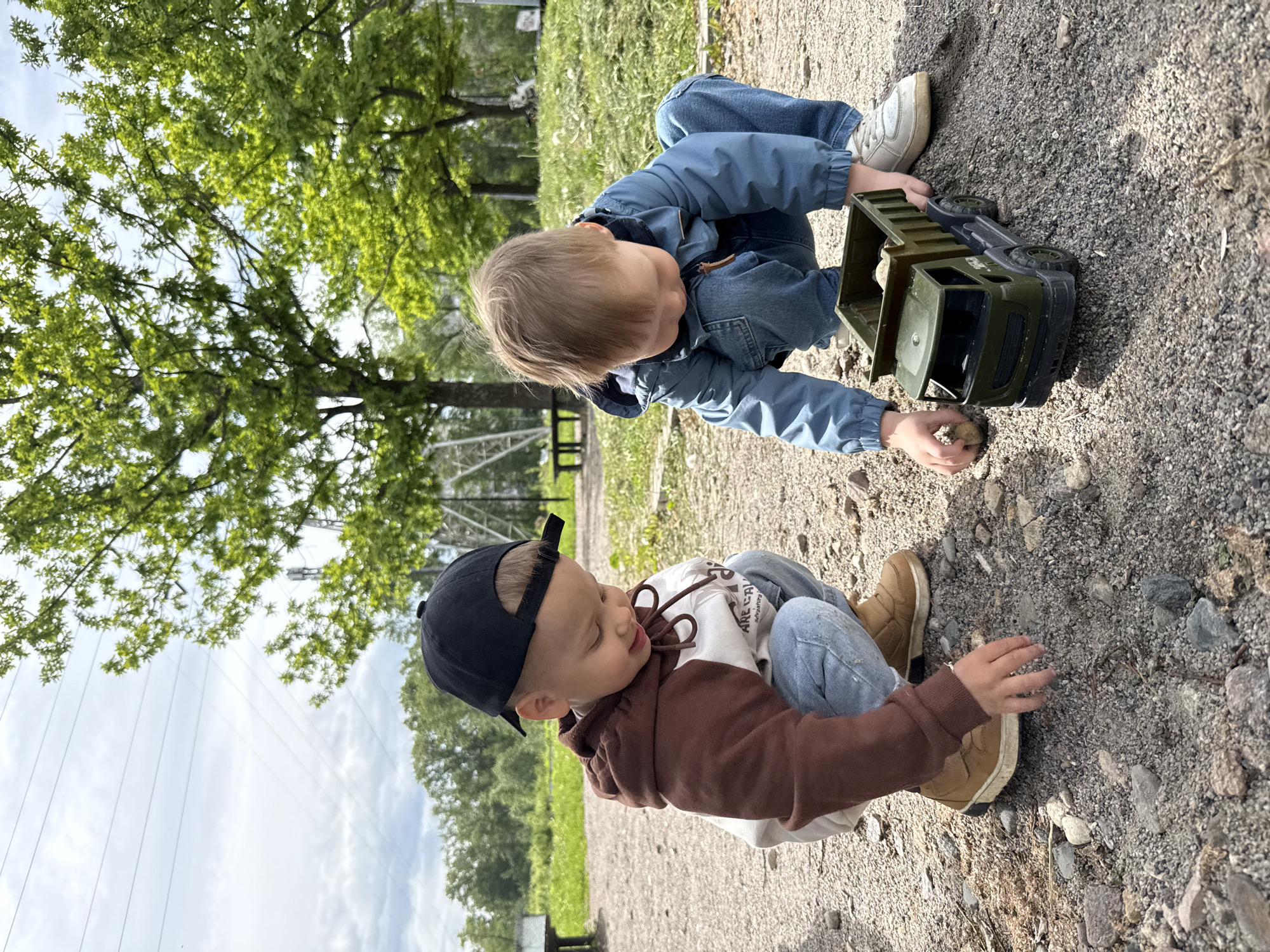 Two young boys are playing on a gravelly ground, presumably outdoors in a park setting, given the presence of trees in the background. The boy closer to the viewer is wearing a brown and white hoodie, jeans, a black baseball cap, and tan boots. He is crouching down, seemingly interacting with the gravel or other small objects on the ground. The other boy is wearing a denim jacket and jeans, with white sneakers. He's leaning over an olive green toy truck, possibly loading it with gravel or dirt. The background includes grass, trees, and a metal structure that may be part of a park or recreational area. The orientation of the image is rotated. No location information is supported.