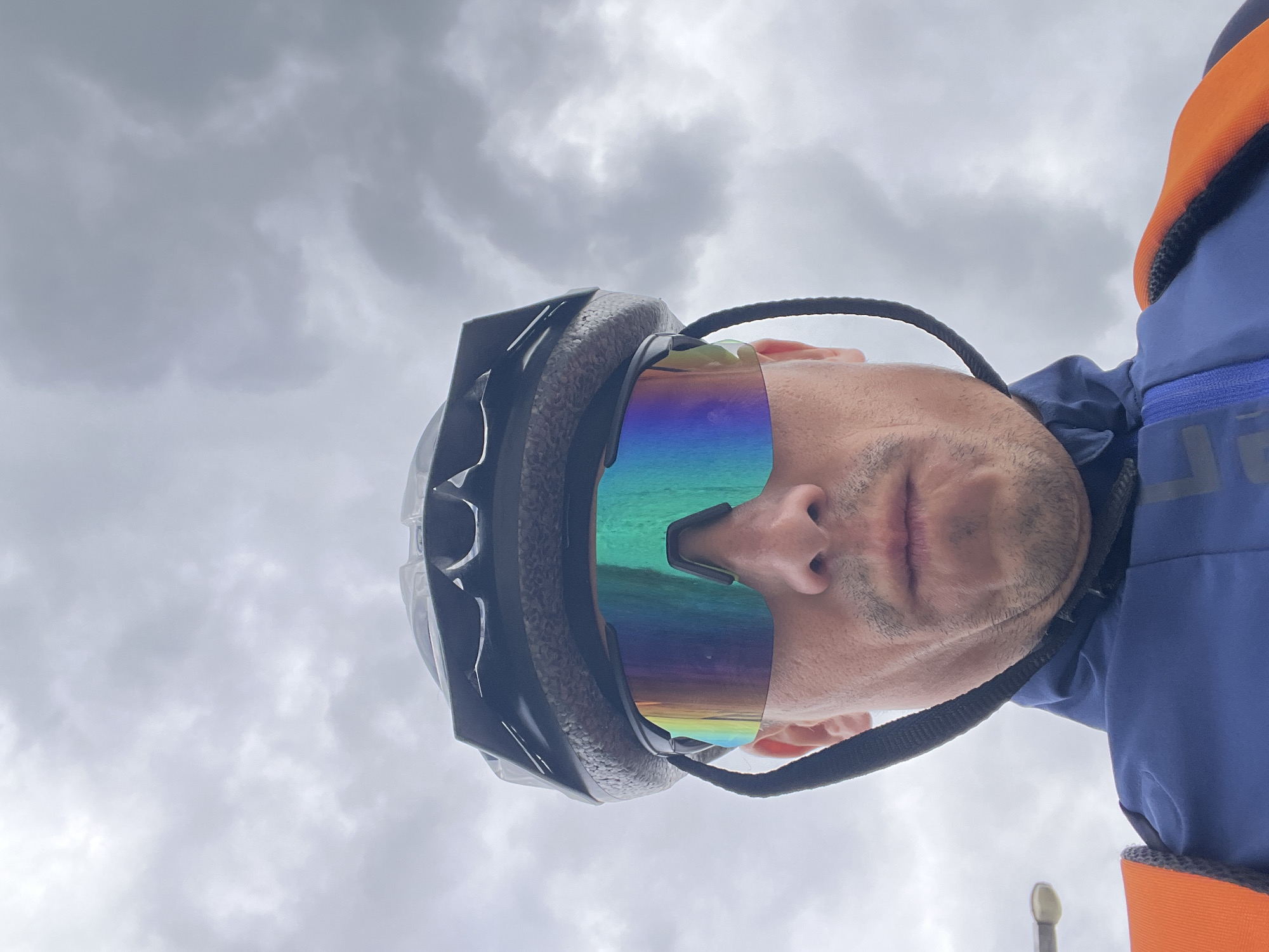 A close-up, low-angle selfie of a man wearing a black cycling helmet and futuristic, iridescent-lensed sunglasses. He is dressed in a dark blue jacket with an orange vest over it. The background consists of a heavily overcast, grey sky. The setting appears to be outdoors, possibly in Chaikovsky, Russia, given the cloudy atmospheric conditions that are common there, particularly during transitional seasons. There are no other people or distinguishing urban features visible in the frame.