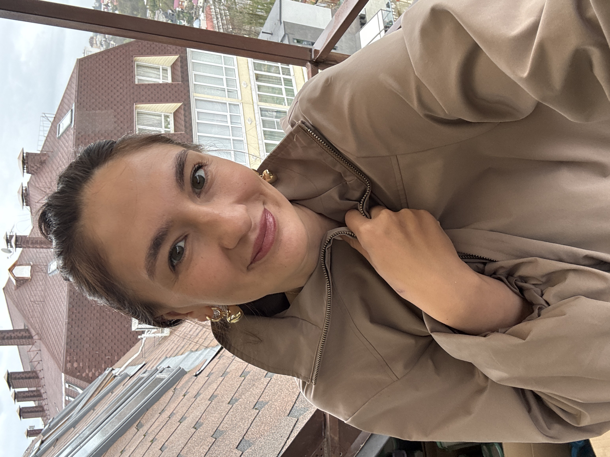 A woman is taking a selfie. She is wearing a beige jacket and gold earrings. The background shows buildings in a city, identifiable as Sochi, Russia due to the architectural style and urban landscape. The woman is standing on a balcony.