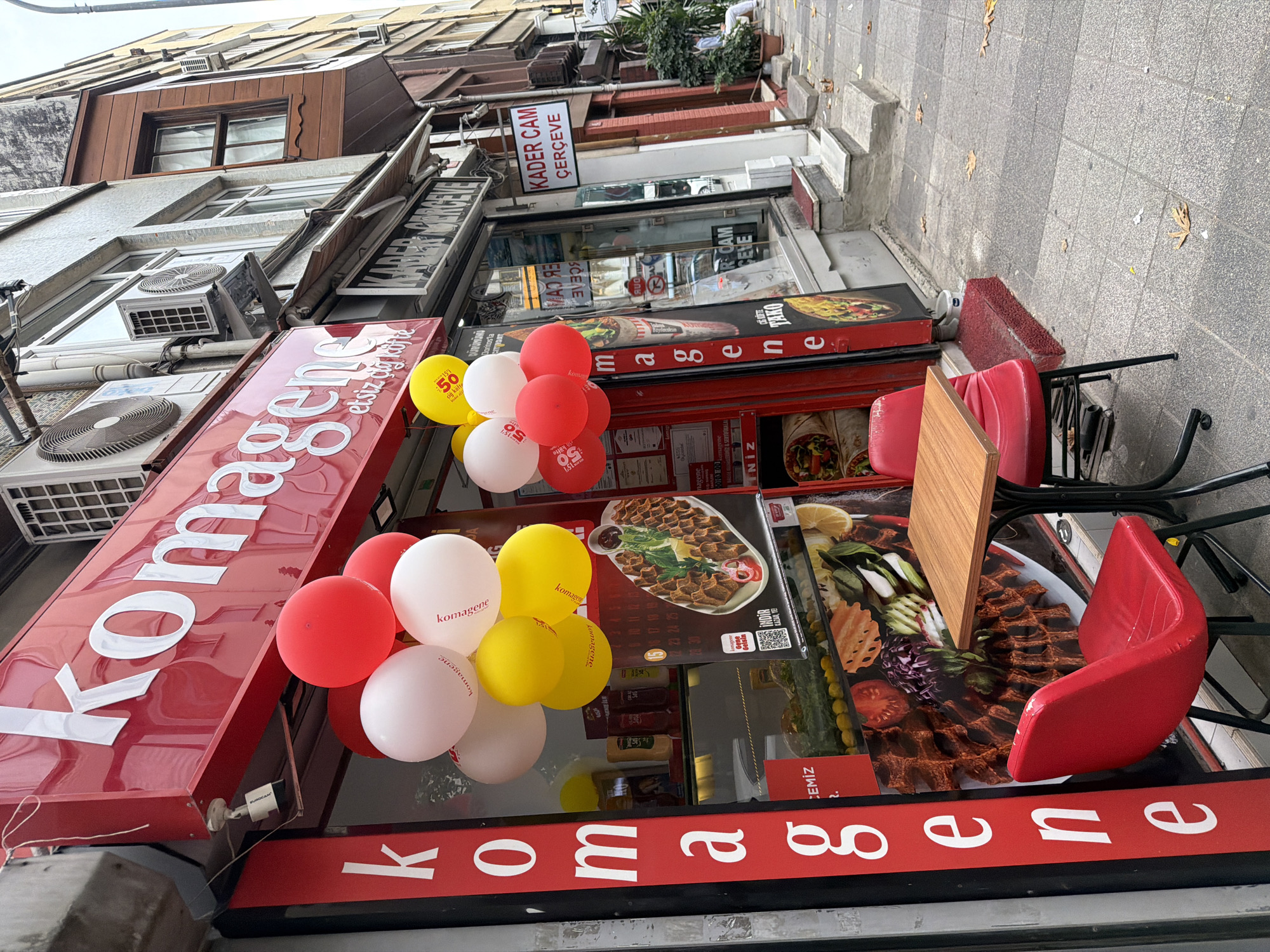 The image depicts the exterior of a "Komagene" restaurant in Kadıköy, Turkey, on a cloudy day. The storefront features a large red sign with white lettering that reads "Komagene," along with the slogan "etsiz çiğ köfte." A vertical red banner on the left side of the storefront also displays the word "Komagene" in white, vertically stacked letters.
In front of the restaurant, a small outdoor seating area is set up with a square wooden table and two red chairs. Several red, white, and yellow balloons are tied to the awning above the entrance, some of which have the "Komagene" logo printed on them.
The shop window displays images of food, including what appears to be çiğ köfte and wraps. Several bottles of sauces are visible on shelves inside. Next door, a sign for "KADER CAM ÇERÇEVE" (Kader Glass Frame) is visible.
The sidewalk in front of the establishments is paved with grey tiles, and fallen leaves are scattered across it, suggesting an autumn season. A person is seated on a ledge across the street, partially visible. The overall environment is that of a commercial street with multiple businesses.
