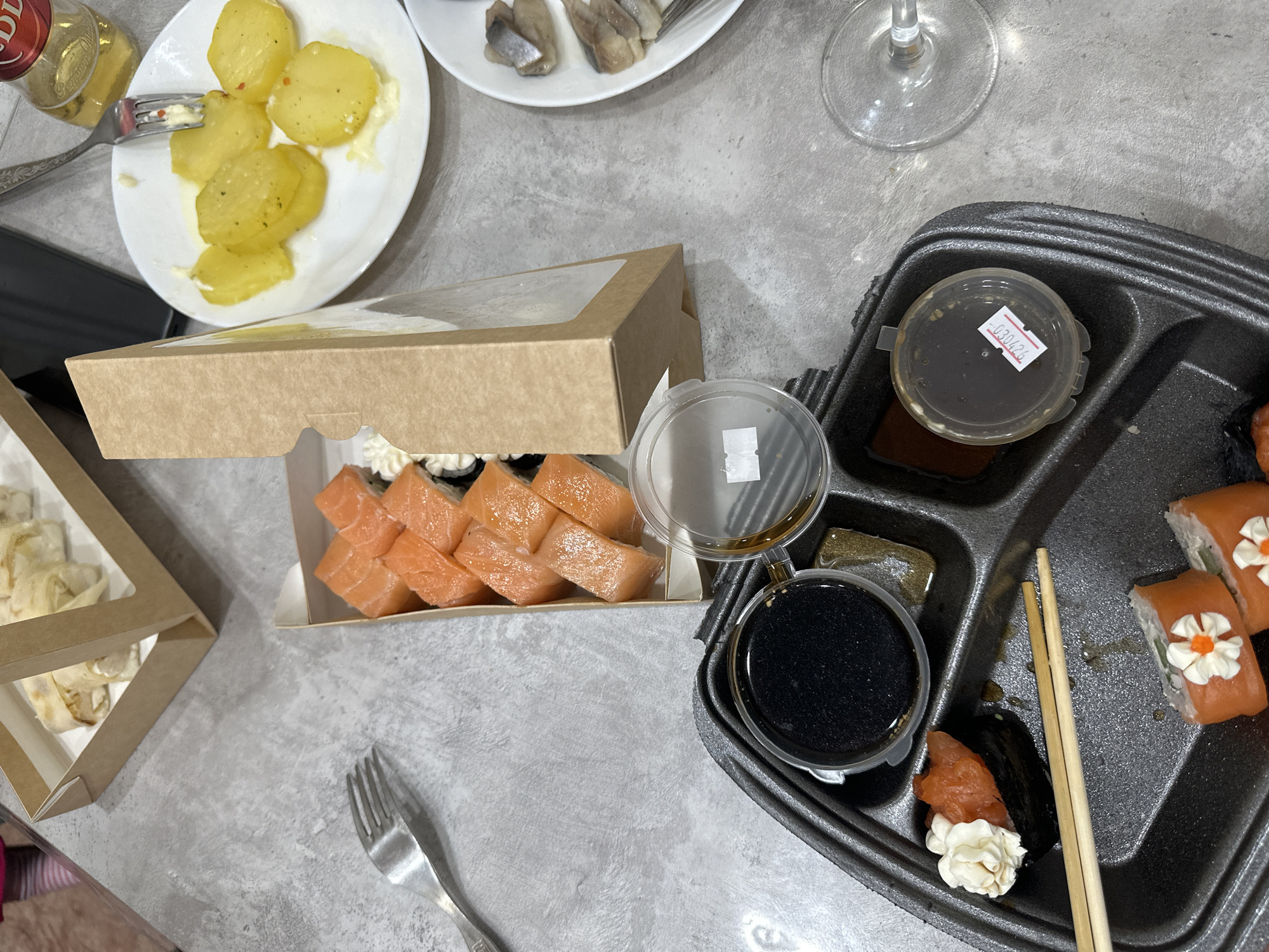 A still life photograph, taken from above, shows a meal laid out on a gray, textured surface. The meal consists of a variety of dishes, including sushi, potatoes, and herring.
A cardboard box containing sushi rolls with salmon and cream cheese is prominent. Adjacent to it is a black plastic tray holding soy sauce containers, chopsticks, and more sushi rolls, some of which are decorated with what appears to be a floral cream cheese design.
To the upper left of the frame, a white plate holds slices of boiled potatoes seasoned with herbs. Above it, another white plate contains pieces of herring. There's also a bottle of oil and a wine glass.
A fork lies on the surface near the sushi boxes. The scene is indoors, likely in a private home. The date code "030426" is visible on one of the sauce containers. The city of Chaikovsky, Russia, is not evident from the image.