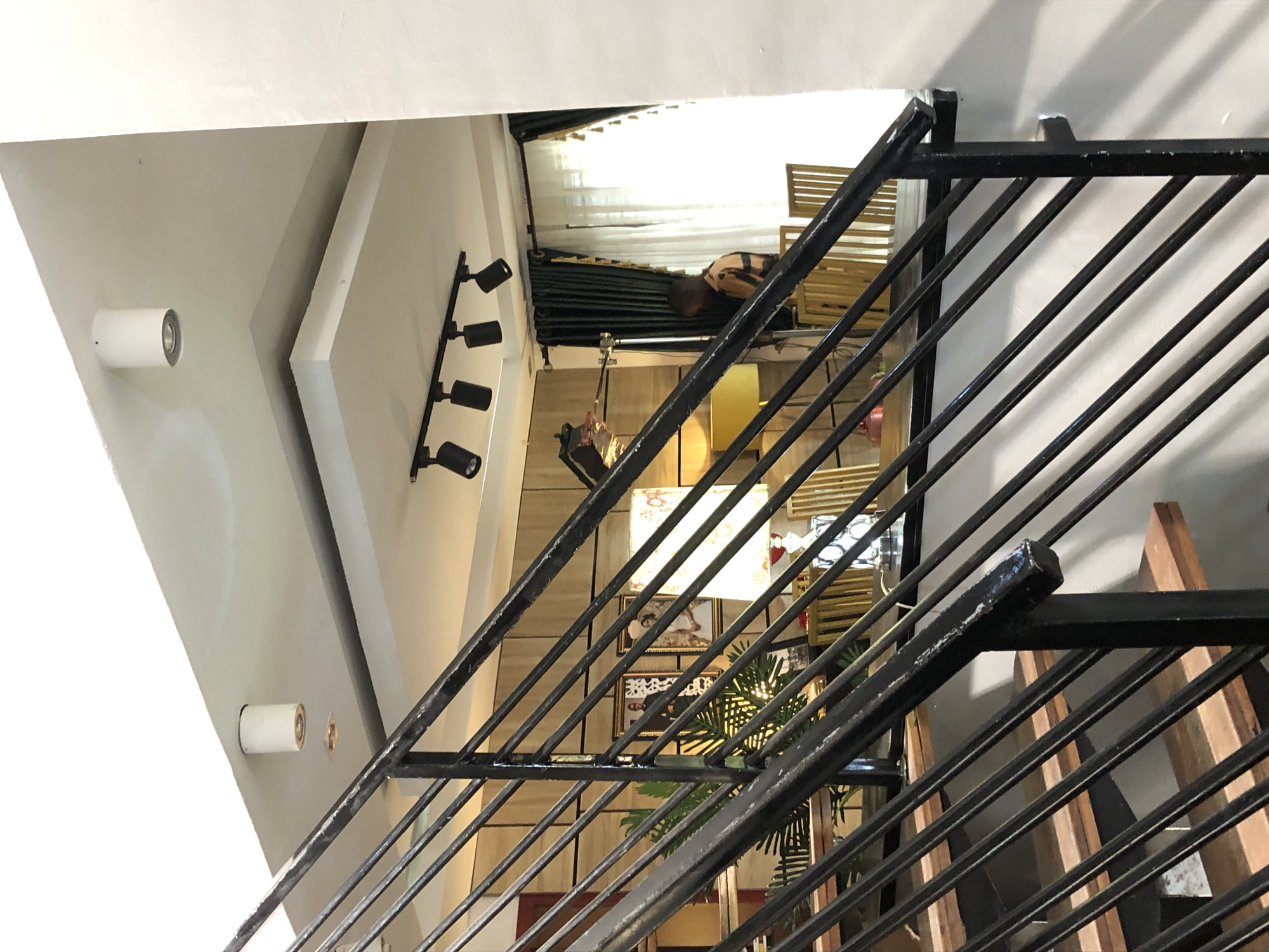 An indoor, multi-level space, possibly a cafe or commercial establishment, is viewed from an elevated perspective looking down. The dominant feature in the foreground is a black metal staircase railing, characterized by numerous diagonal bars, extending from the bottom right upwards towards the left. Wooden stair treads are partially visible behind this railing.
On the lower level, situated below and to the left of the staircase, a person is seated or reclining on furniture made of light wooden slats, possibly a bench or part of a booth. The individual has dark hair and is wearing a striped top. Behind them, windows are covered by light-colored curtains.
The walls and ceiling are white or light-toned. Modern lighting fixtures are visible: a track light system with four black, cylindrical spotlights is mounted on the left wall, pointing downwards. Two white, cylindrical wall-mounted lights are also present on different wall sections. Decorative elements on the lower level include framed pictures or art on the wall, a green, palm-like plant, and a patterned lampshade. The scene is well-lit, conveying a contemporary interior design. There is no visible text.