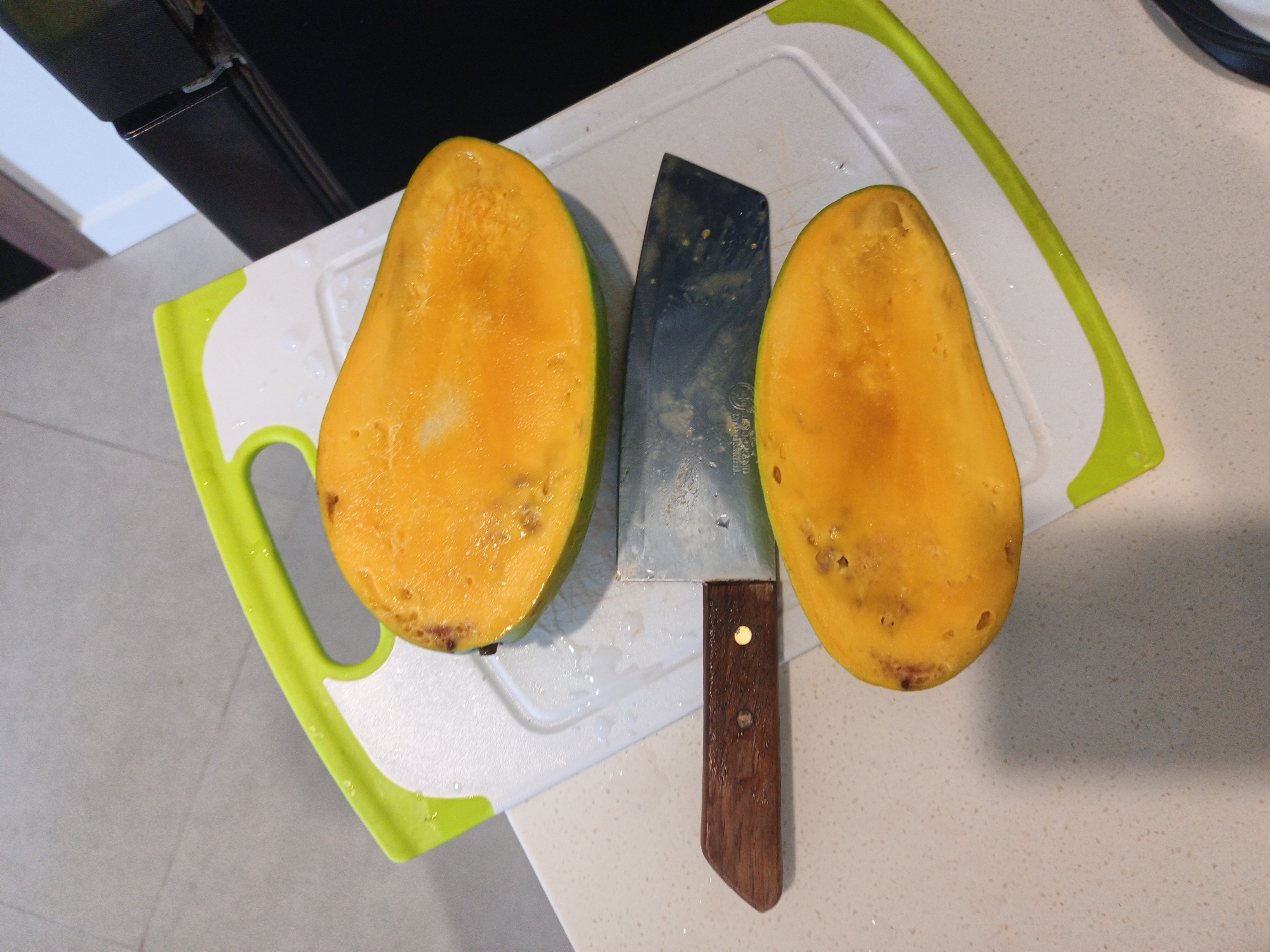 In Bac Nha Trang Ward, Vietnam, two halves of a ripe mango, showcasing their bright orange flesh, rest on a white cutting board with a green edge. The mangoes appear to have been recently sliced, as evidenced by the glistening moisture on the cutting board. Positioned between the two fruit halves is a cleaver-style knife with a stainless steel blade and a dark wooden handle. The knife's blade bears faint speckles. The cutting board is situated on a light-colored countertop. In the upper portion of the image, a dark, rectangular object, possibly a kitchen appliance, is partially visible. The overall scene depicts food preparation in a domestic setting.