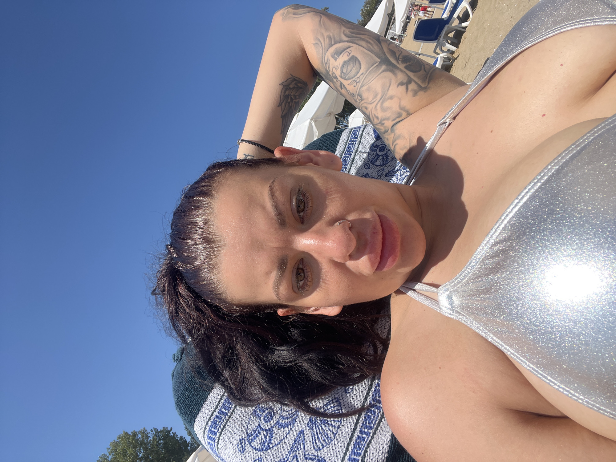 A woman with dark hair pulled back in a ponytail and tattoos on her arms lies on a striped blanket on a beach. She is wearing a silver bikini. The background shows a clear blue sky, beach chairs, umbrellas, and other beachgoers. The sun is shining brightly. The location is Sozopol, Bulgaria.
