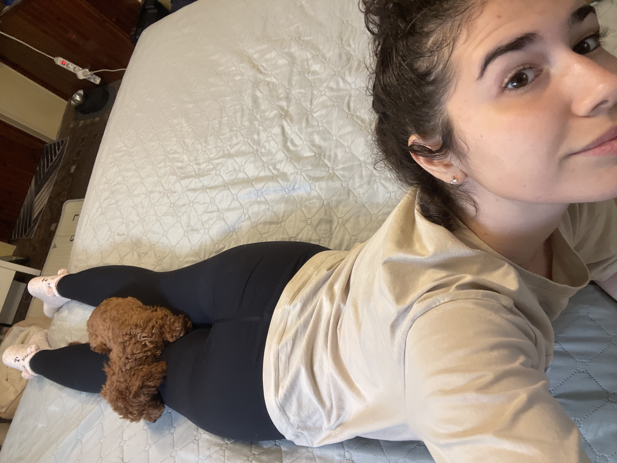 A woman lies on a bed with a light blue quilted comforter. She is wearing black leggings, pink socks, and a light beige long-sleeve shirt. A small, brown poodle sits between her legs. The scene appears to be indoors in a bedroom setting. The location in Sofia, Bulgaria cannot be determined from the image.