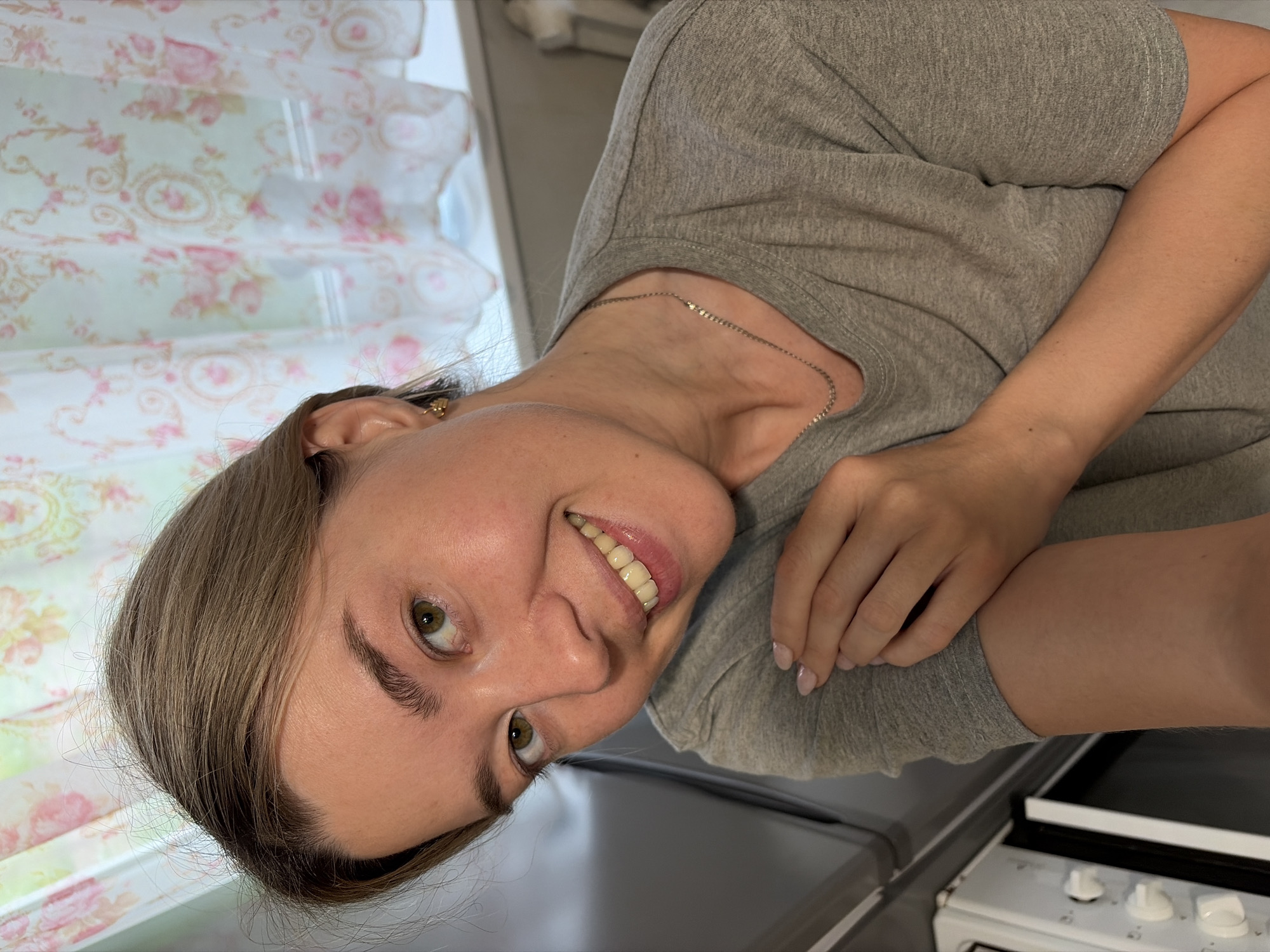 A woman with light brown hair and green eyes is indoors, partially visible. She is wearing a gray t-shirt and a silver chain necklace. Her left ear has a gold earring. She is smiling slightly. Behind her is a window with a floral patterned curtain. A portion of a stove is visible at the bottom right of the frame. The exact location in Chaikovsky, Russia cannot be determined from the image.