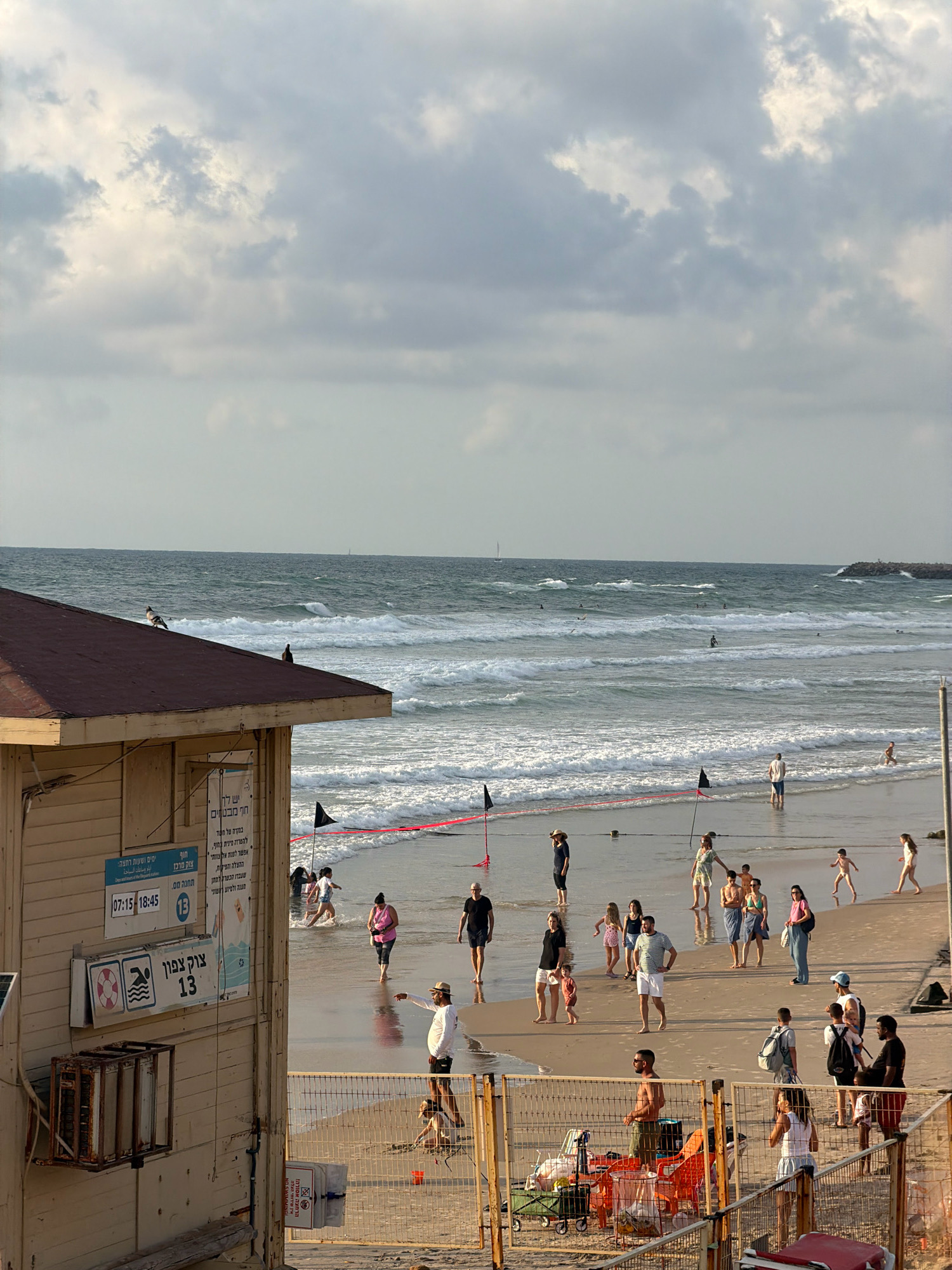 This is a medium-angle, eye-level shot of a crowded beach in Herzliya, Israel, likely in the late afternoon based on the lighting. The foreground is dominated by a weathered, wooden lifeguard station with a dark brown roof. Signs with Hebrew text and icons are affixed to its side. Two pigeons are perched on the roof of the station.
The middle ground features a sandy beach where numerous people are scattered. Some are walking along the shoreline, while others are wading in the ocean. Several black flags on poles are planted in the sand, marking a boundary. The ocean is moderately rough with white-capped waves rolling towards the shore. A red rope demarcates a section of the beach, possibly for swimming. A few distant figures are visible in the water, some possibly surfing.
The background shows the expanse of the sea meeting a hazy sky filled with dramatic clouds, suggesting an approaching change in weather or simply a cloudy day. A distant breakwater or pier is visible on the right side of the frame. The overall impression is one of a lively public space with people enjoying the beach despite the overcast conditions.