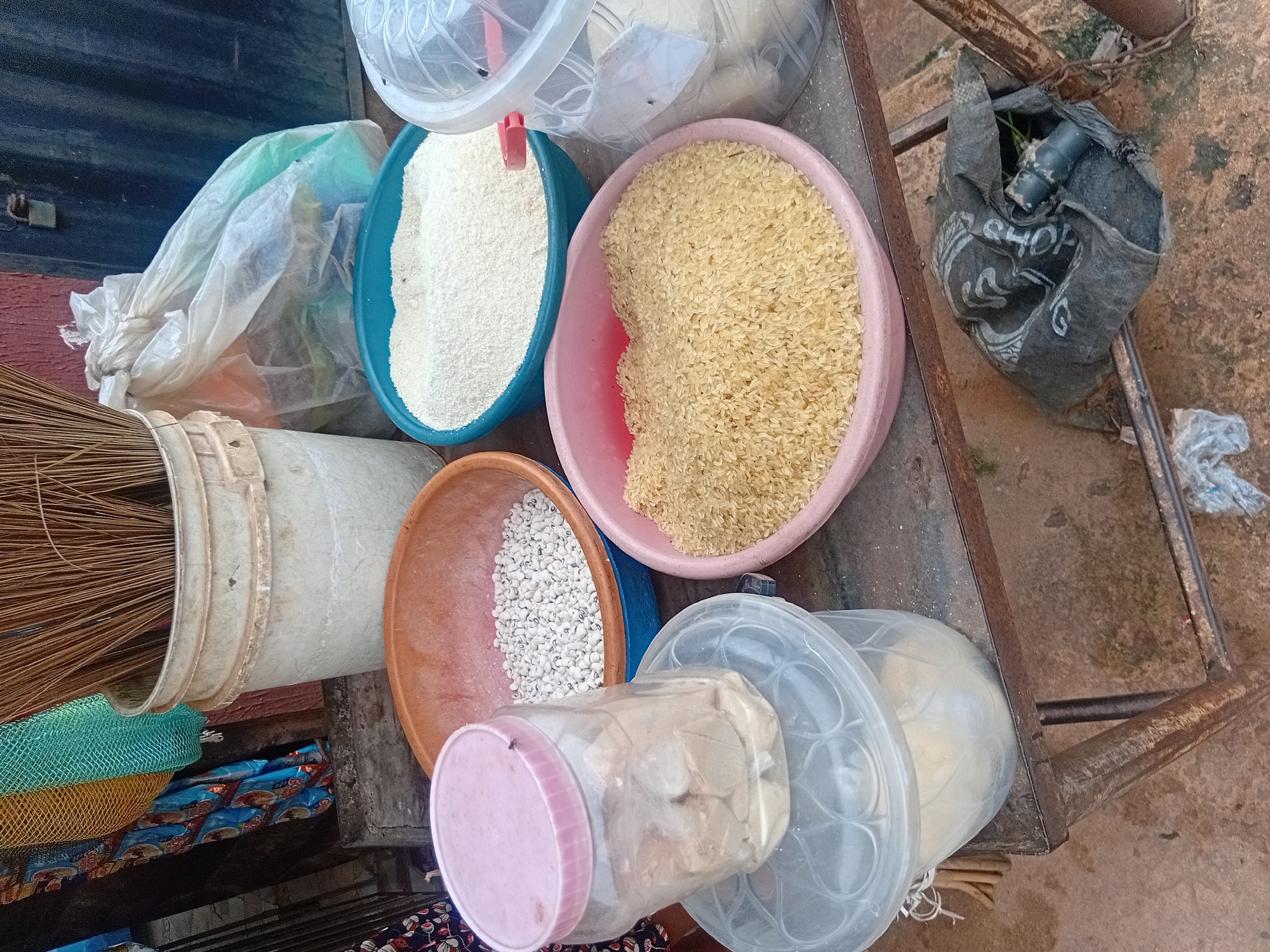 The image depicts an outdoor market stall in Nkpor, Nigeria, featuring a collection of food items and household goods. In the foreground, a metal table holds various containers. A large pink bowl contains a generous amount of uncooked yellow rice. To its left, a transparent jar with a pink lid holds what appears to be dried yams or cassava, with a fly resting on the lid. Beneath the jar, a clear plastic container with a patterned lid is partially visible.
Behind these items, a light orange bowl is filled with white beans, possibly black-eyed peas. Further back, a turquoise bowl is filled with white rice or a similar grain. To the right of the turquoise bowl, a partially visible clear plastic container holds more white grain.
In the background, a tall white bucket is filled with a bundle of dry broomsticks. Next to it, a large, crinkled white plastic bag contains colorful items, possibly textiles or more goods. The stall is situated on a dirt ground, with a rusty metal frame supporting the table and a chain to its right. In the far background, a dark green corrugated metal door with a padlock is visible. The overall scene suggests a typical Nigerian marketplace offering a variety of dry goods for sale.