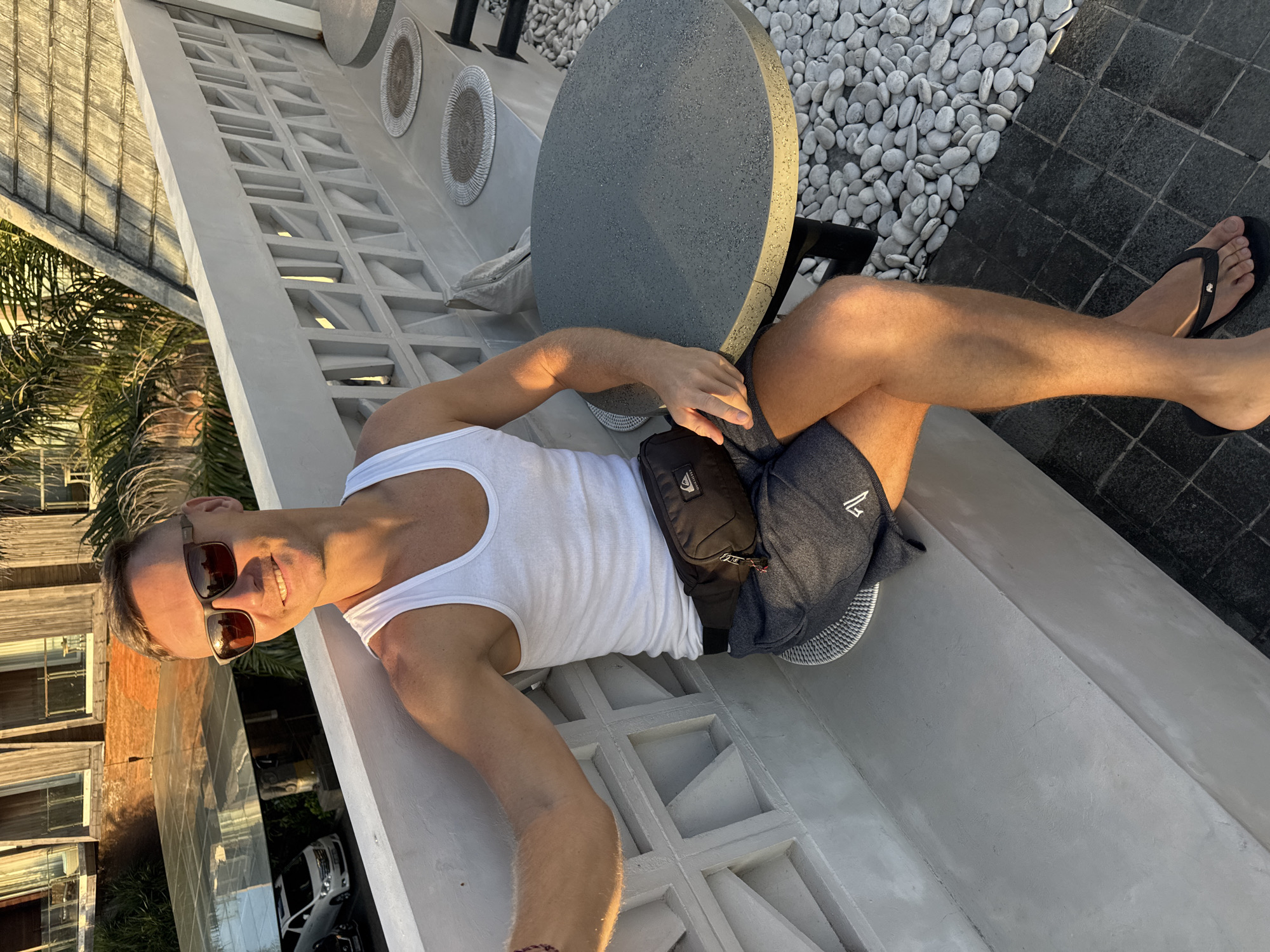 The photograph captures a man reclining on a white concrete ledge adjacent to a round grey table in what appears to be an outdoor patio setting. He is dressed in a white tank top, dark shorts, and black flip-flops and has a black waist bag slung across his chest. He is also wearing sunglasses. The man is facing the camera with a slight smile.
The background features white pebbles near the table and a building facade with windows, suggesting an urban environment. Palm trees can be seen in the distance. The distinct architectural style and general ambiance indicate that the location might be in Kuta, Indonesia.