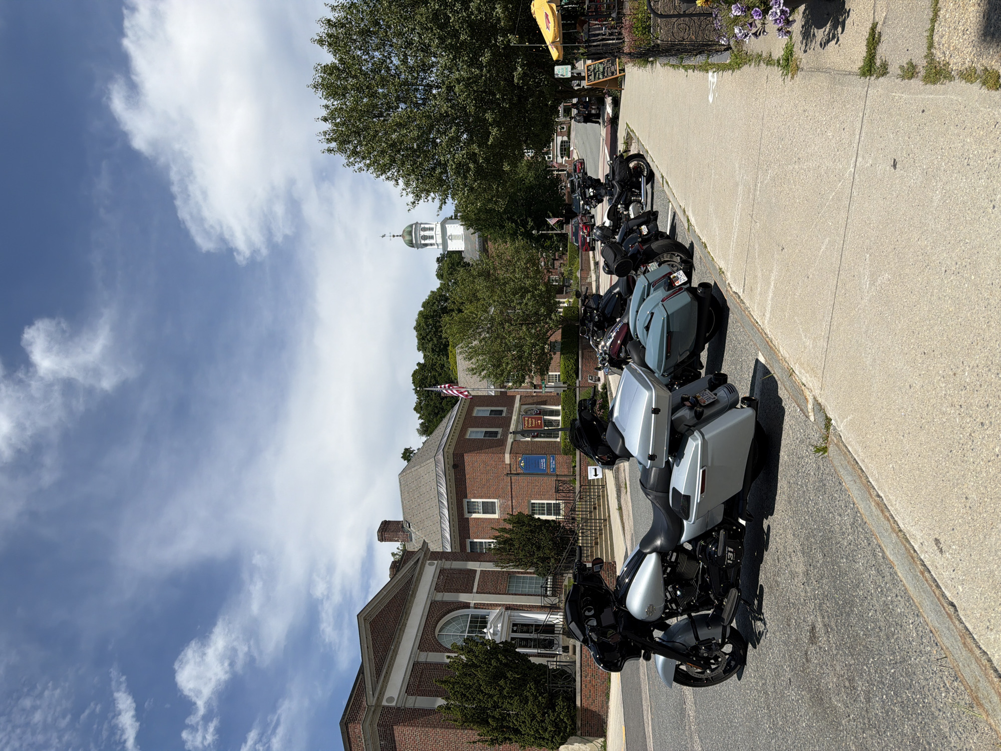 The image depicts a street scene in Peterborough, United States of America, on a bright, cloudy day. In the foreground, several motorcycles are parked along the side of the road. The closest motorcycle is a silver Harley-Davidson, featuring saddlebags and a large front fairing. Behind it, other motorcycles of varying colors are visible, including a dark grey one and several black ones.
The street runs alongside a sidewalk, which has a black metal fence bordering a garden area with purple flowers. A yellow umbrella is set up over a table at what appears to be an outdoor seating area for a cafe or restaurant. A chalkboard sign stands near the entrance to this area.
Across the street, there are several brick buildings. The building on the left has a grand entrance with an arched window and classical architectural details. To its right, another brick building with a gabled roof is visible. Further in the background, a white building with a green steeple and clock tower stands against the sky, which is filled with scattered white clouds against a blue backdrop. Trees are interspersed throughout the scene, with one large tree prominently featured on the right side of the image. An American flag is flying from a flagpole in front of one of the buildings. A few people are casually walking on the sidewalk in the background.
