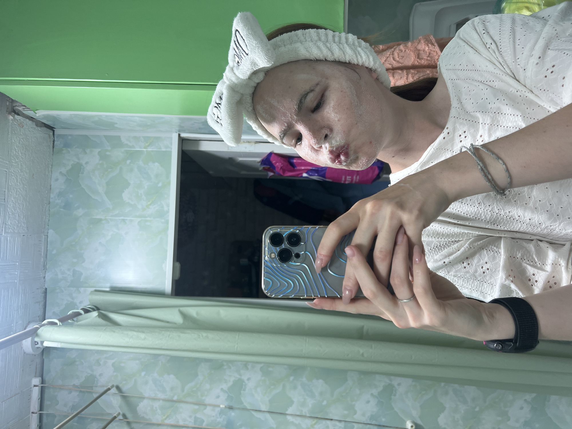 A woman with white foam on her face is taking a selfie with a silver phone. She wears a white top and a white headband with bunny ears. She has a black watch on her left wrist and a silver bracelet on her right. She is pouting her lips and looking at the camera. The photo appears to be taken in a bathroom with light green walls and a green curtain. The exact location in Chaikovsky, Russia cannot be determined from the image.