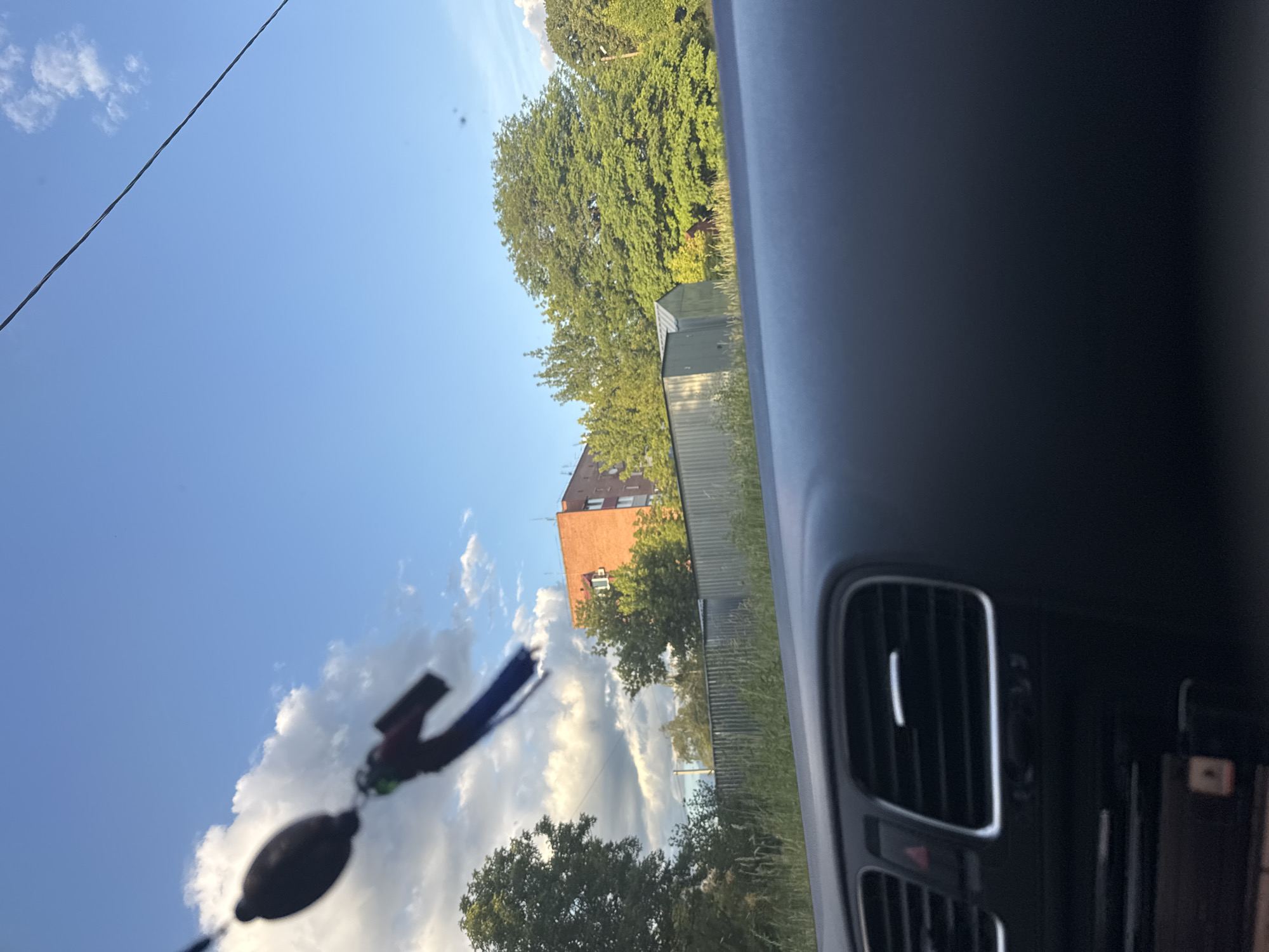 The image, taken from inside a vehicle, shows a view through the windshield of a suburban or rural area. In the foreground, the dashboard of a car is visible, including air vents. Outside, a blue sky with scattered clouds is visible. In the mid-ground, there is a red brick apartment building, a corrugated metal fence, and various trees and greenery. A wire stretches diagonally across the sky. A dark oval object with a blue strap hangs in the upper left of the frame, obscuring part of the sky. The scene appears to be in daylight, likely late afternoon given the warm lighting. There are no visible people or animals. The setting suggests a residential area adjacent to undeveloped land.