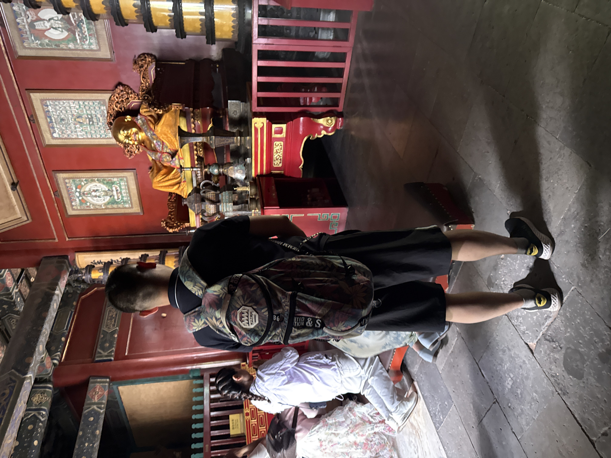 The image shows the interior of a temple in Dongcheng District, China. In the foreground, a person wearing a black t-shirt and black shorts with a floral-patterned backpack stands facing away from the camera, looking towards a gilded statue of a seated Buddha. The statue is adorned with a golden robe and sits on an ornate, red and gold throne. Behind the statue, the temple walls are painted red, and several framed artworks hang on them. To the right of the statue, a tall, multi-tiered yellow and black structure is visible. In the middle ground, to the left of the central figure, a young person with braided hair, wearing a white shirt and white pants, is kneeling on a red cushion. The floor is paved with large, grey stone tiles. The environment appears to be dimly lit, with light sources casting shadows.